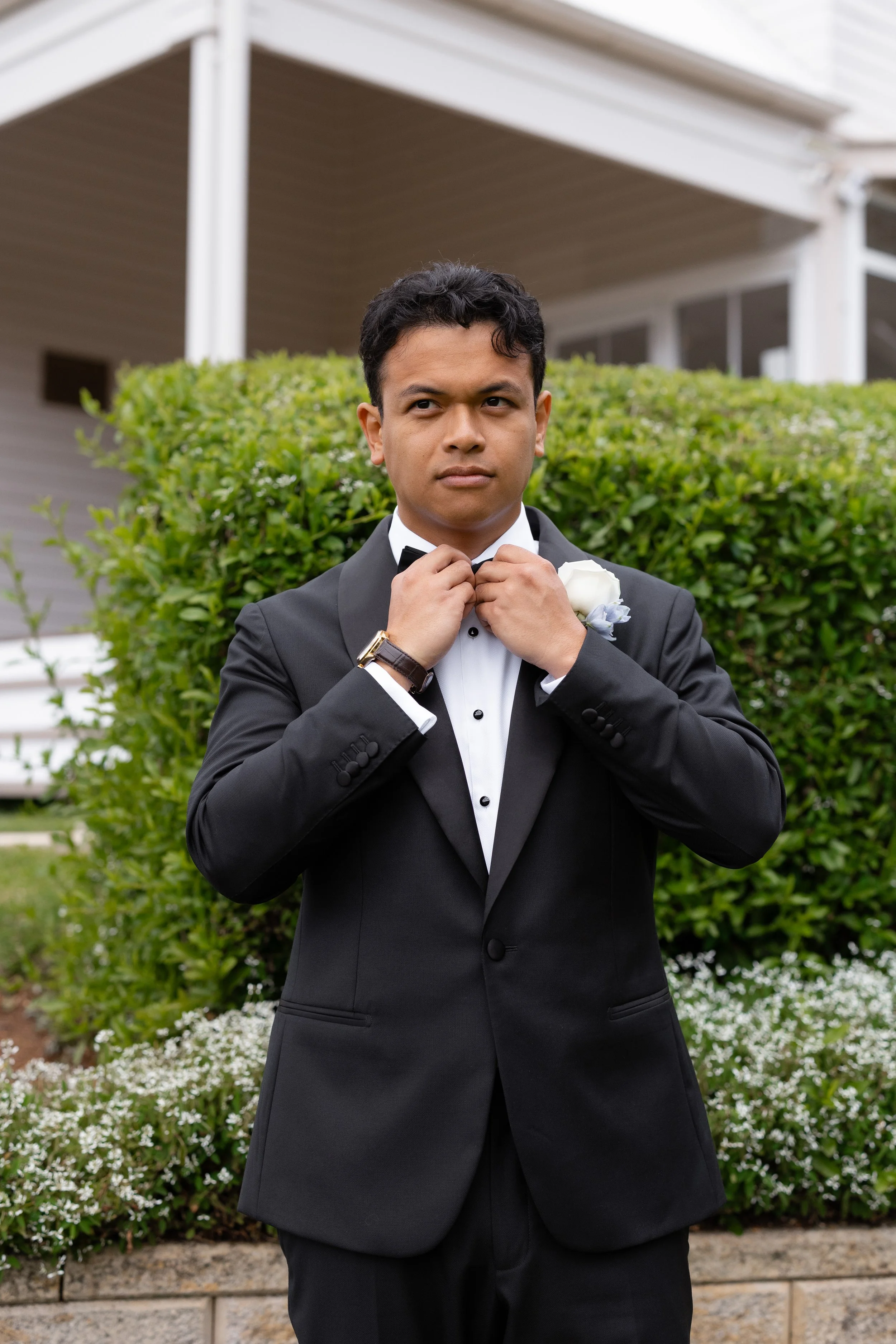 Alicia and Ezra's wedding day: Ezra in a black tuxedo adjusting his bow tie in front of a hedge
