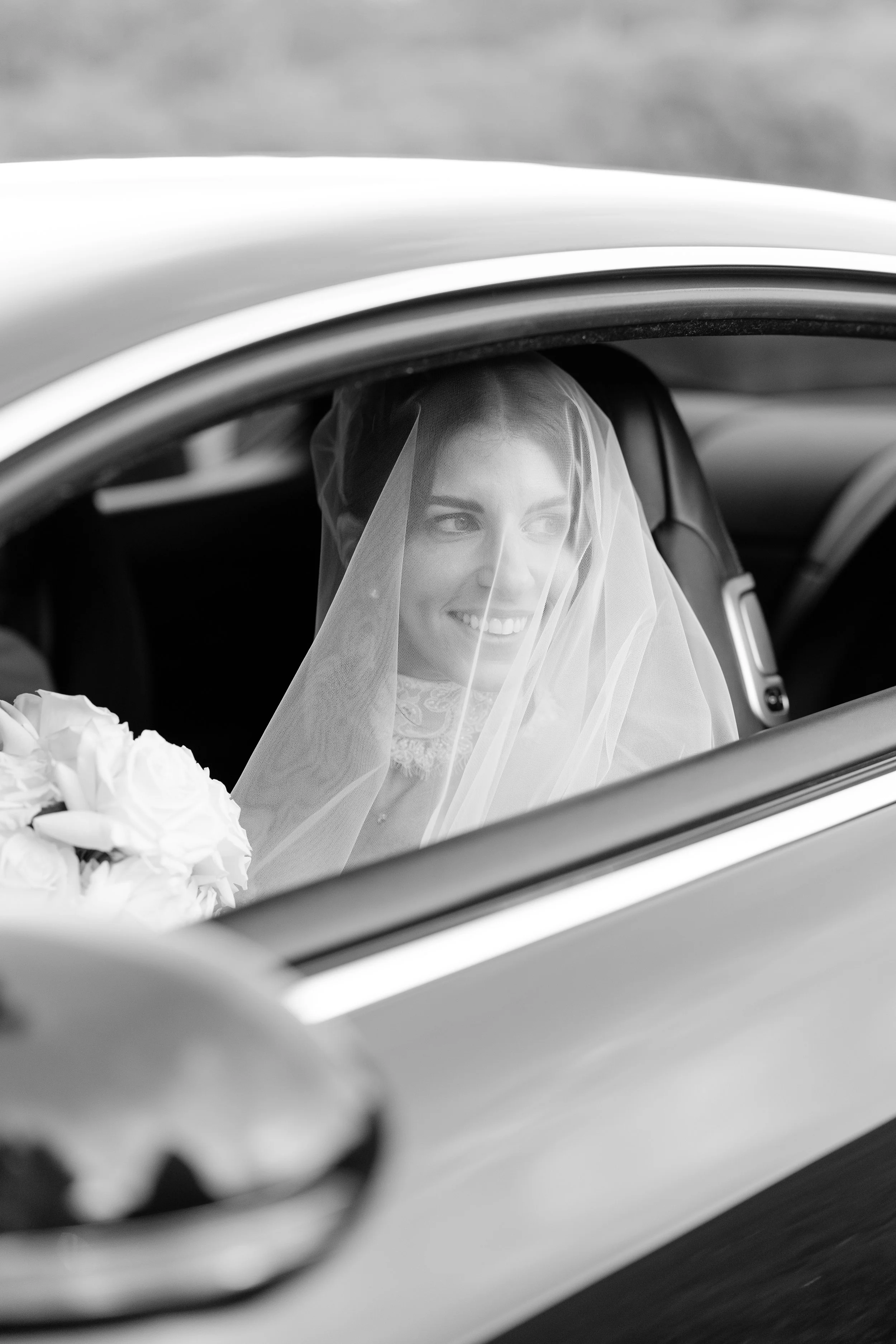 Steph in her veil holding a white bouquet, seen through a car window, in black and white