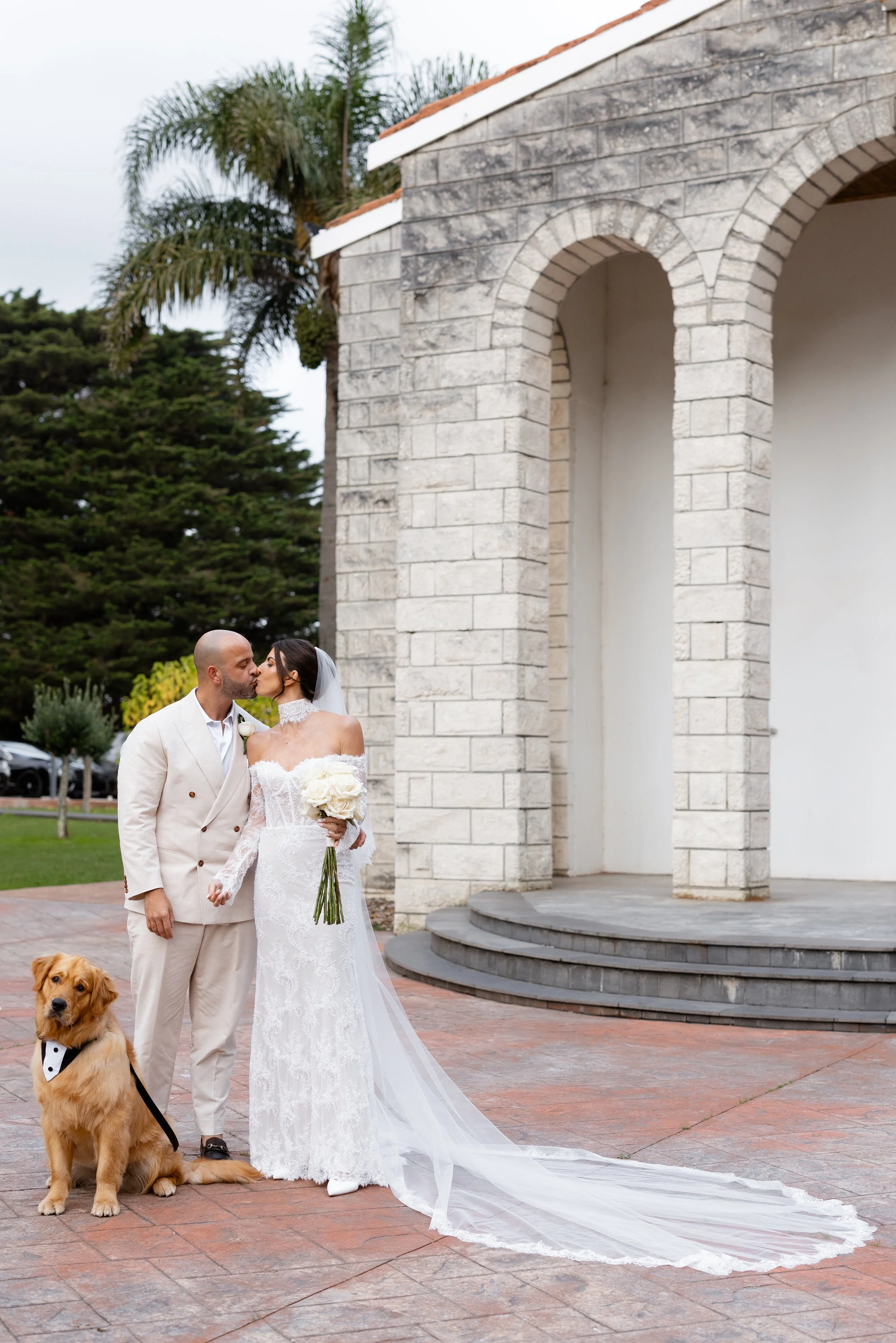 Steph and Alex with their dog beside the stone church arches, her long veil trailing