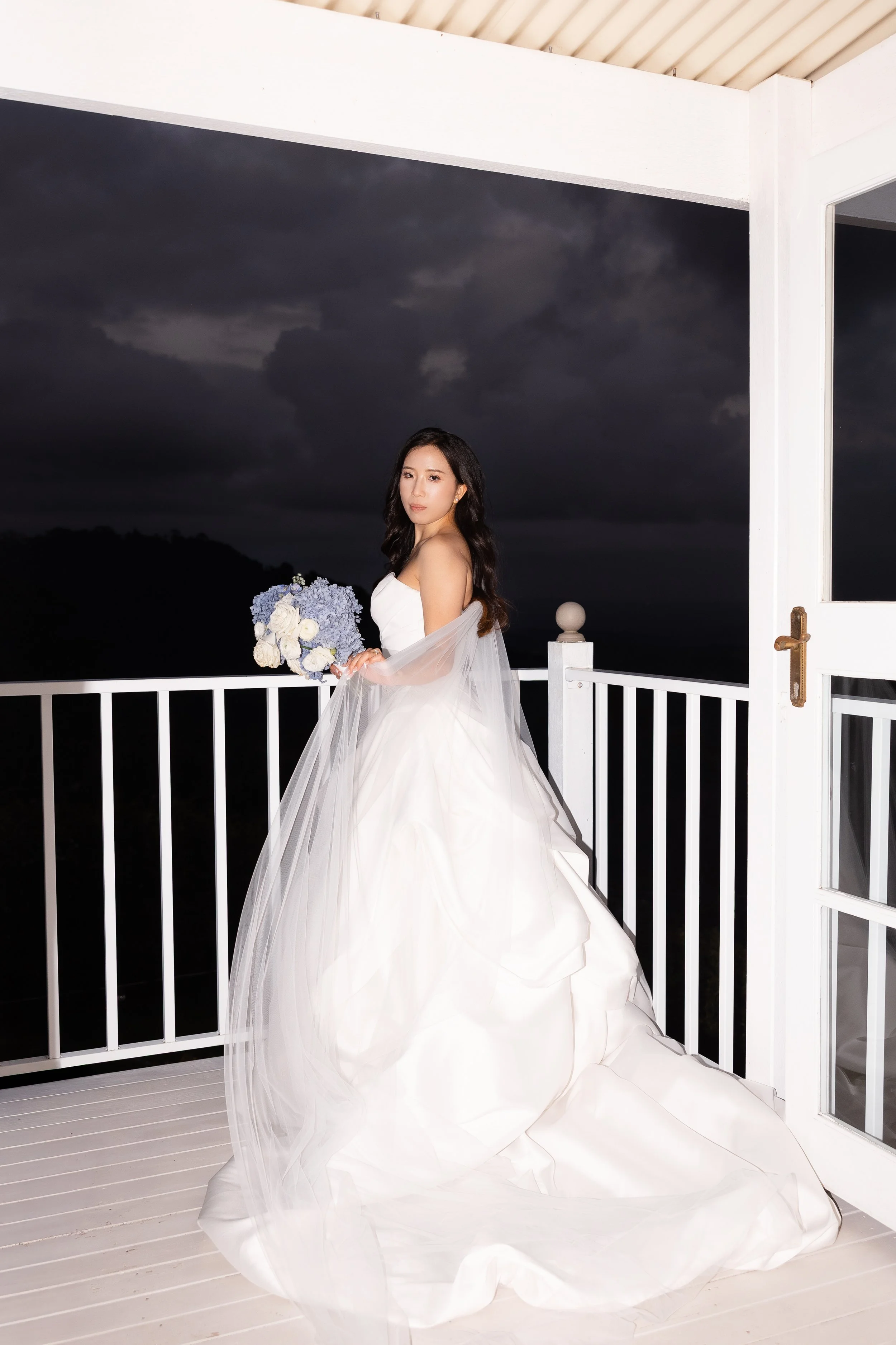 Alicia in her white gown holding a bouquet on a white balcony railing at dusk