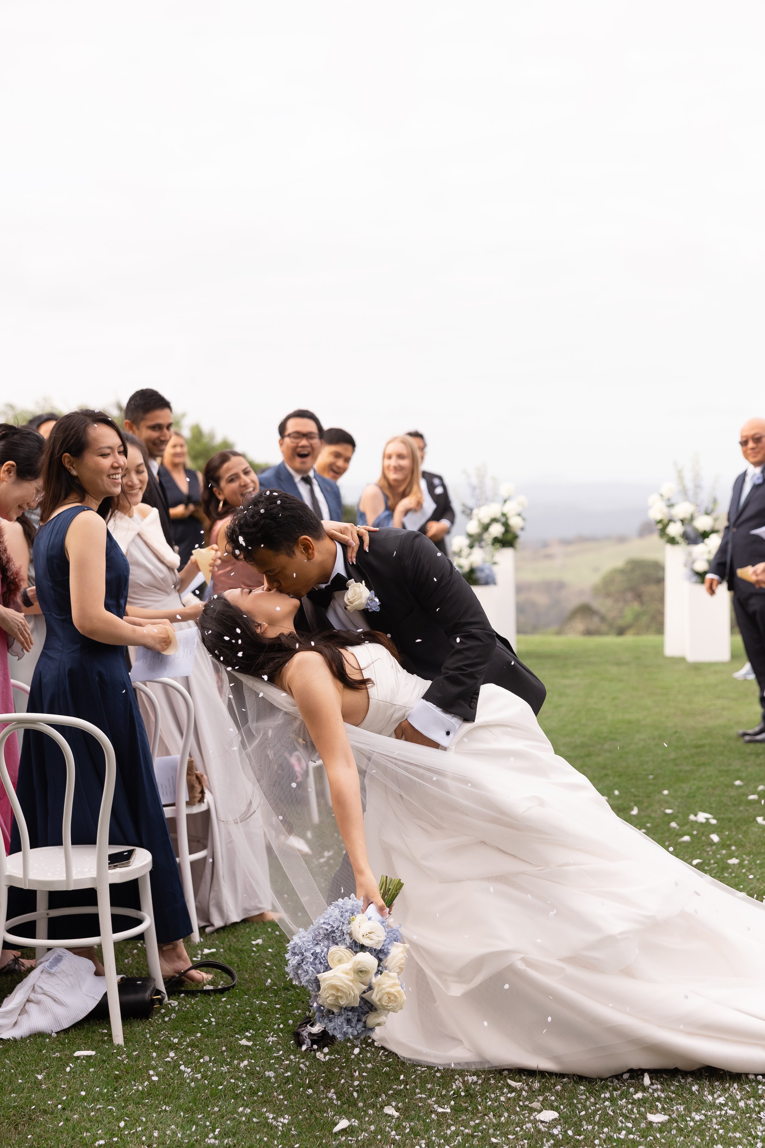 Ezra dipping Alicia for a kiss on the lawn while guests watch from white chairs