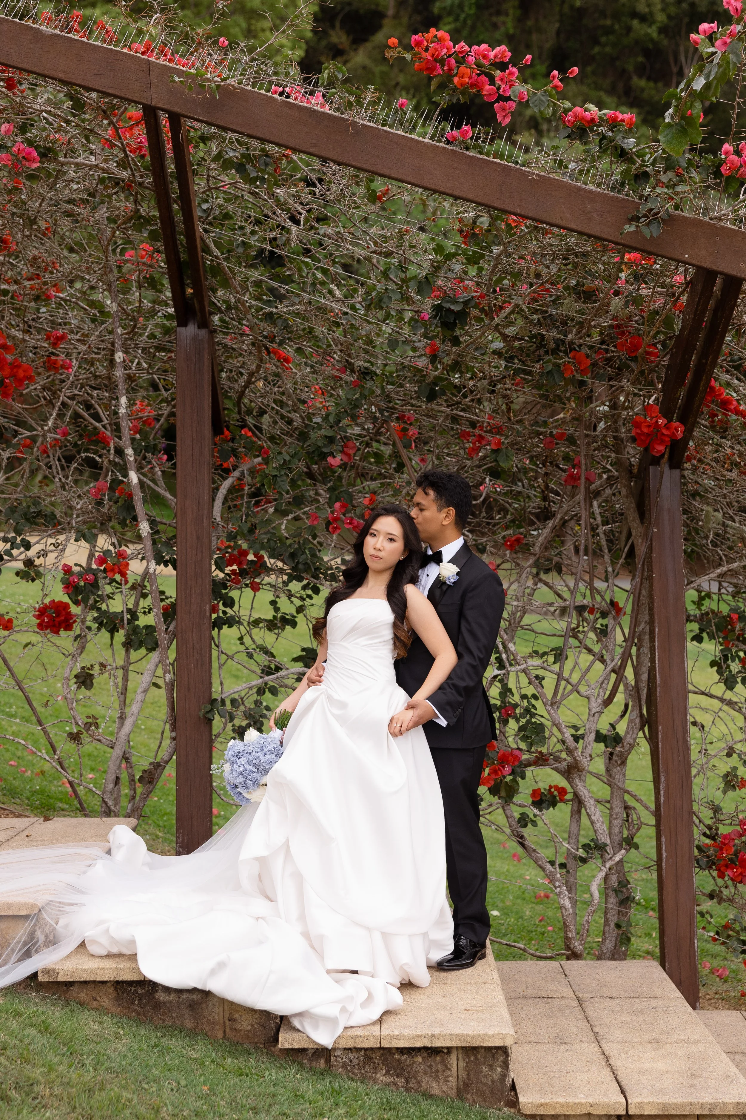 Alicia and Ezra standing together on a wooden platform beneath a rose-covered pergola