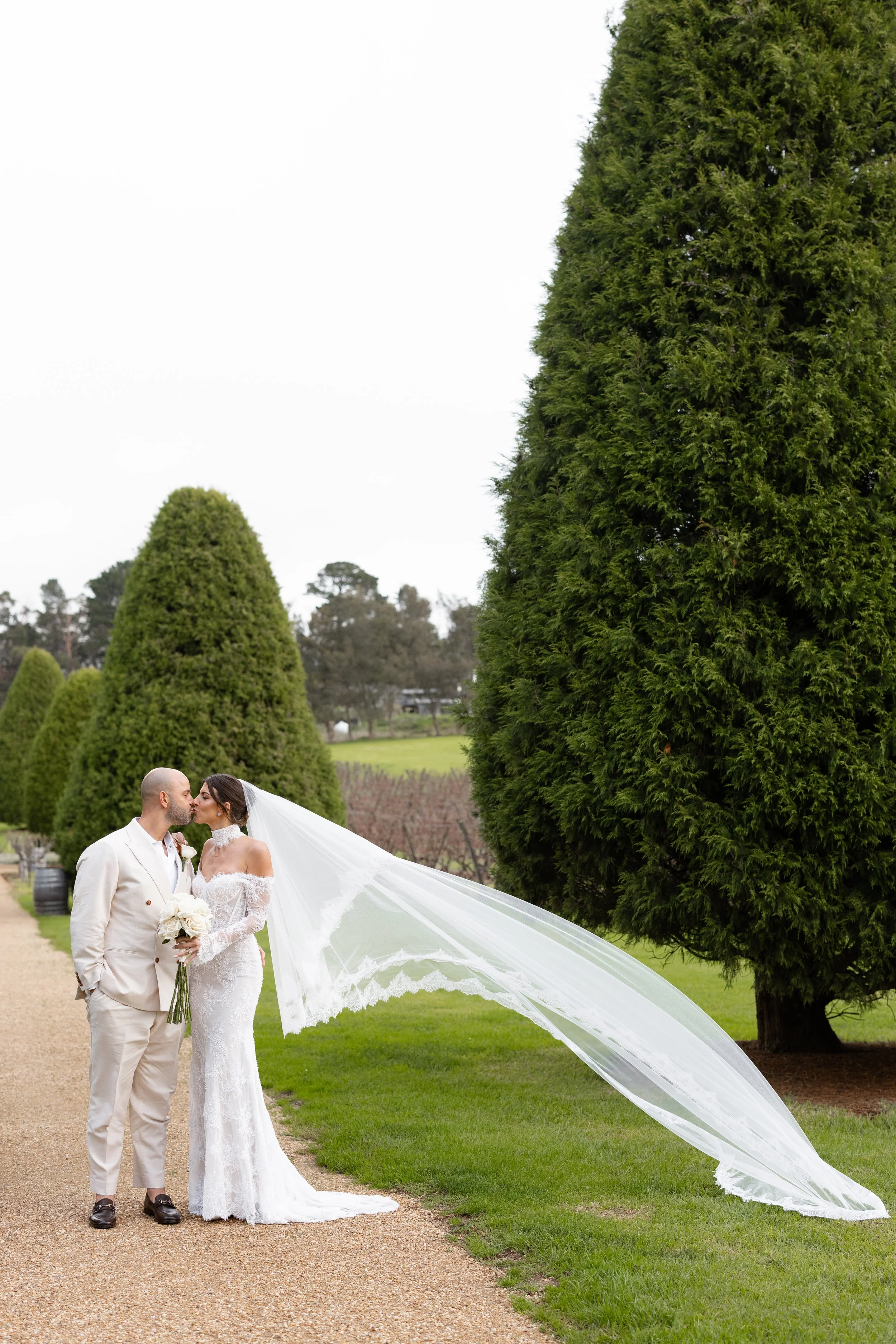 Steph and Alex walk the hedge-lined path at Lindenderry Red Hill, her veil streaming behind