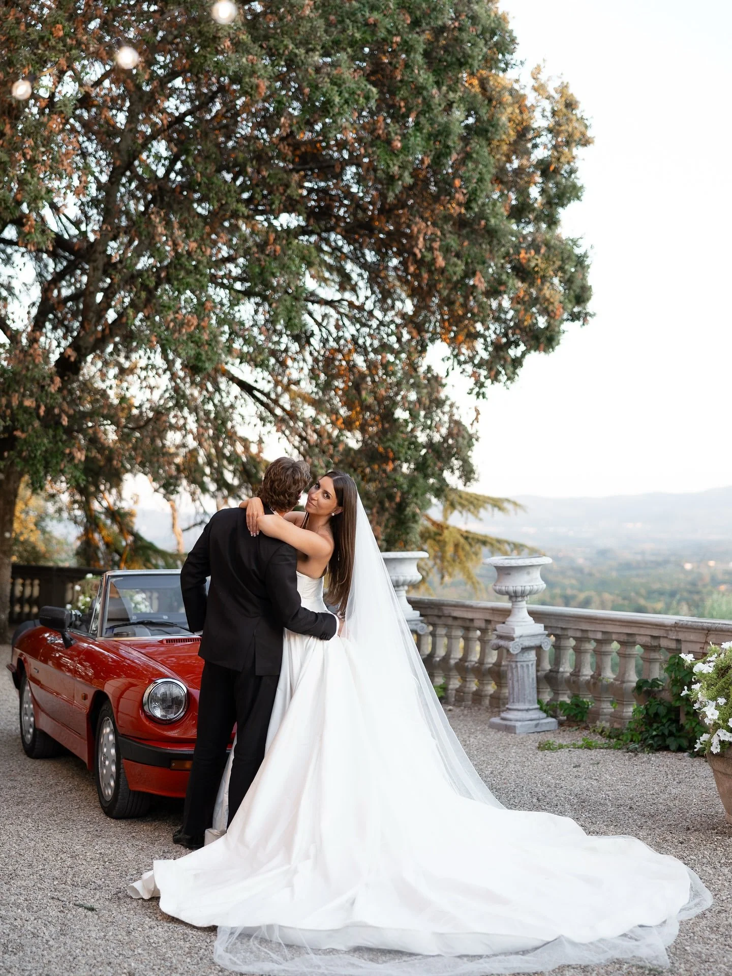 Groom in a black suit embracing bride Marilena in her gown and veil beside a red vintage convertible at Villa Belvedere, Loro Ciuffenna, Tuscany