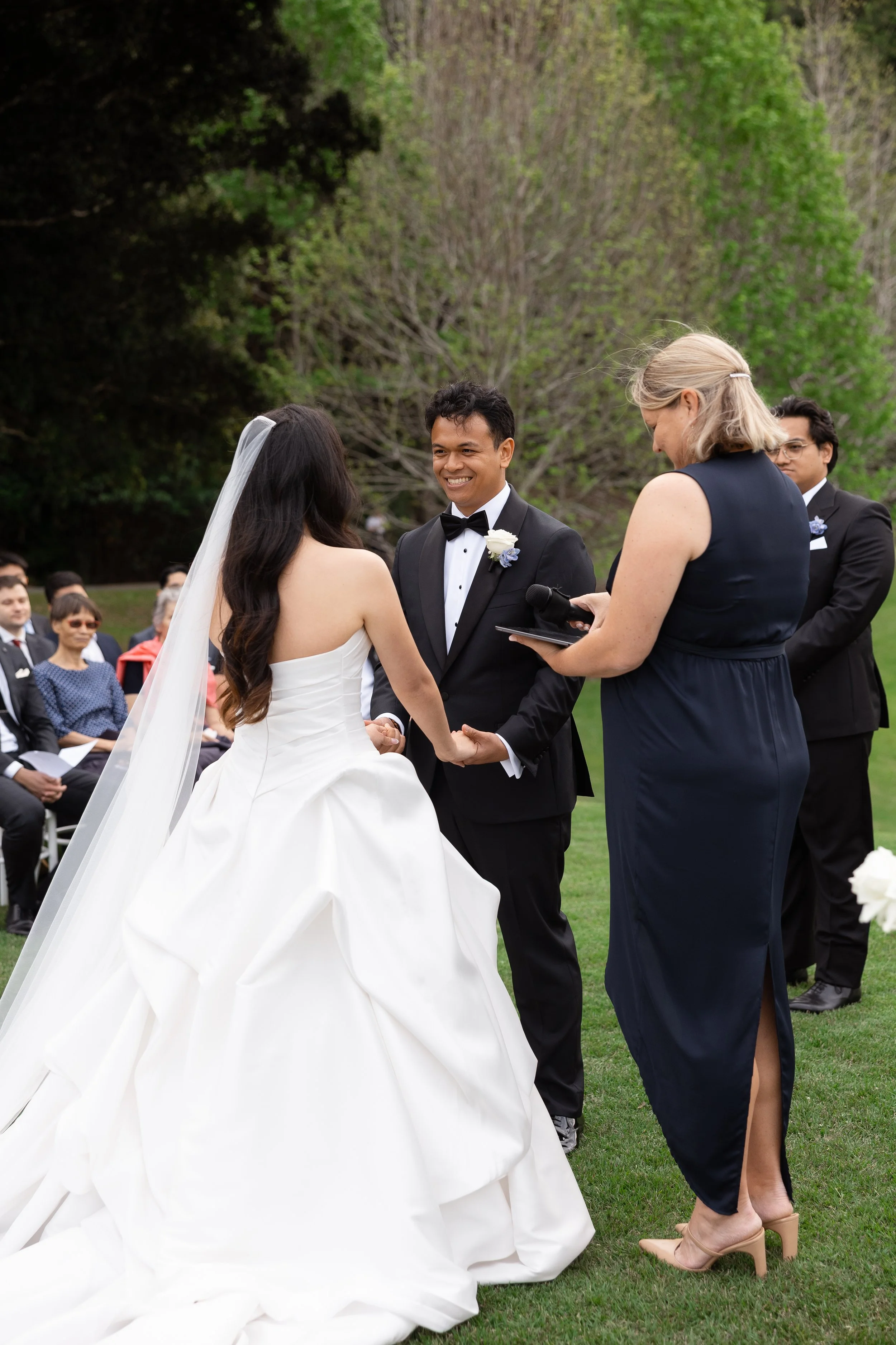Alicia and Ezra holding hands in front of the celebrant in a navy dress during the outdoor ceremony