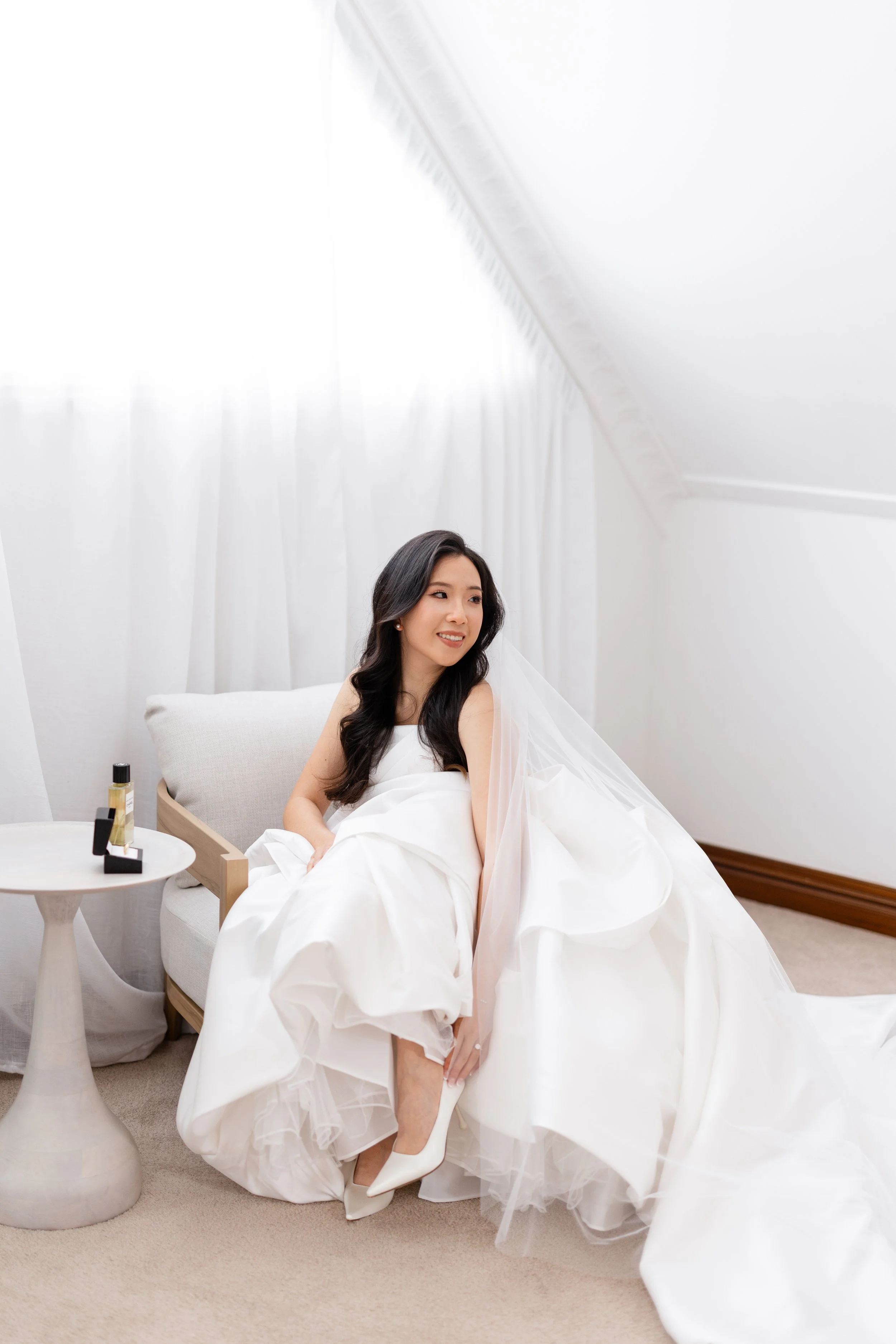 Alicia in her white gown seated in a bright room, touching her white heel
