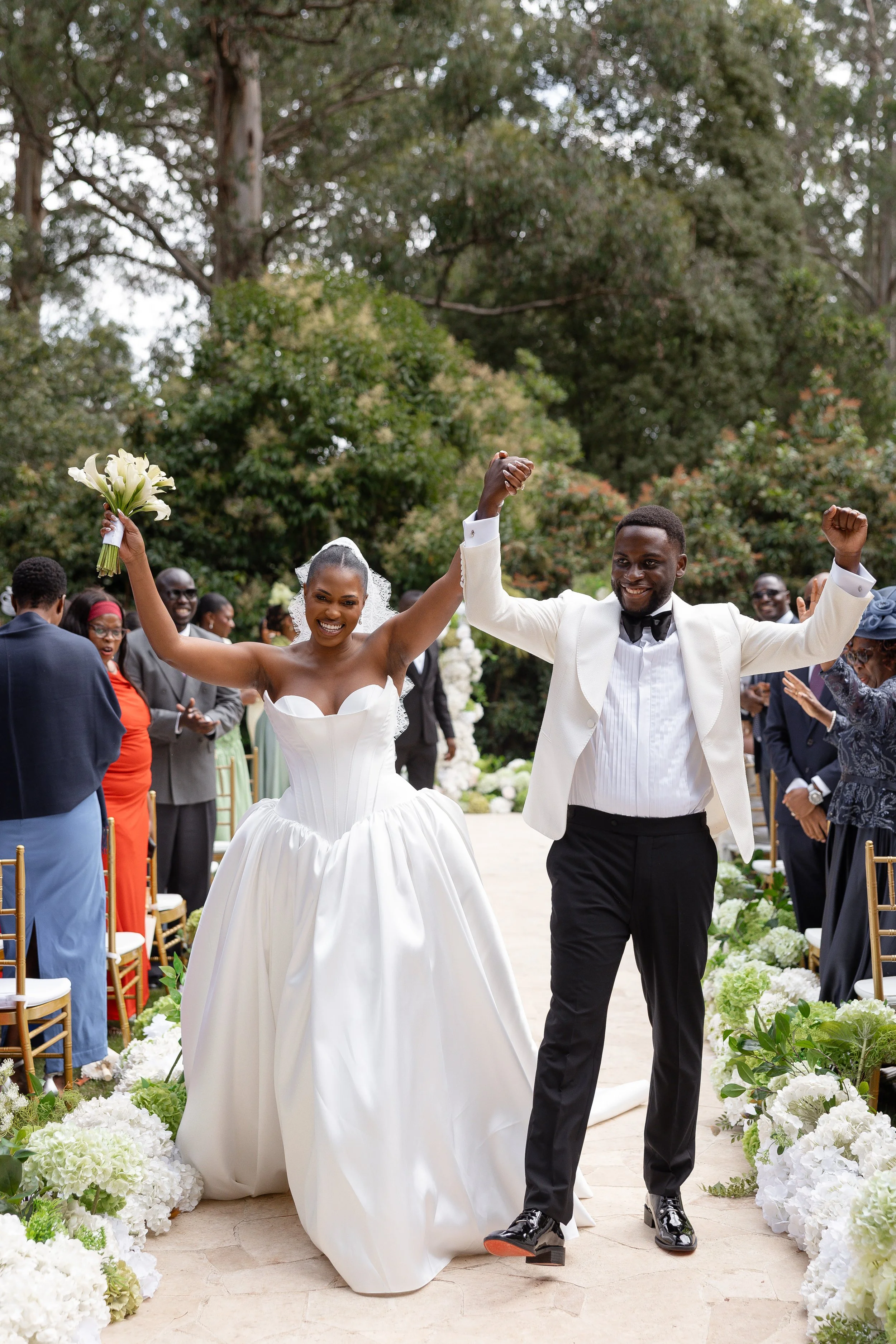 A newly married couple celebrating outdoors, walking down the aisle with arms raised in victory, surrounded by friends and family, with flowers lining the walkway.