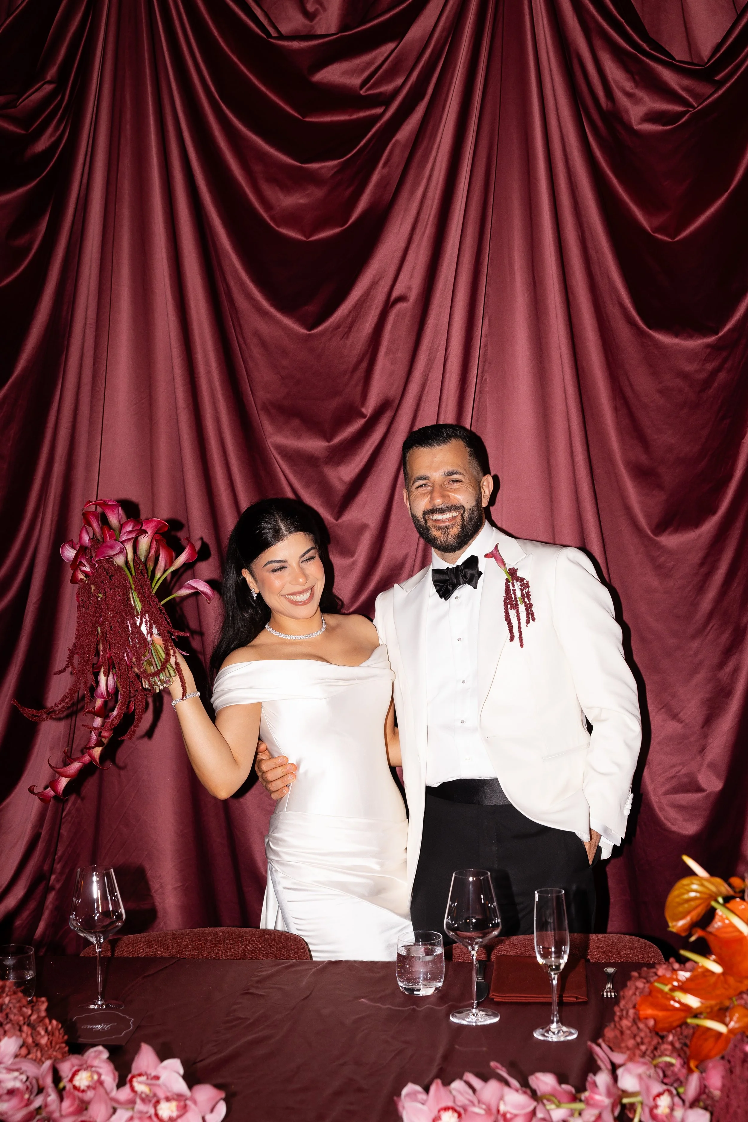 Jihan and Rob in front of burgundy silk drapes, Jihan holding a burgundy bouquet