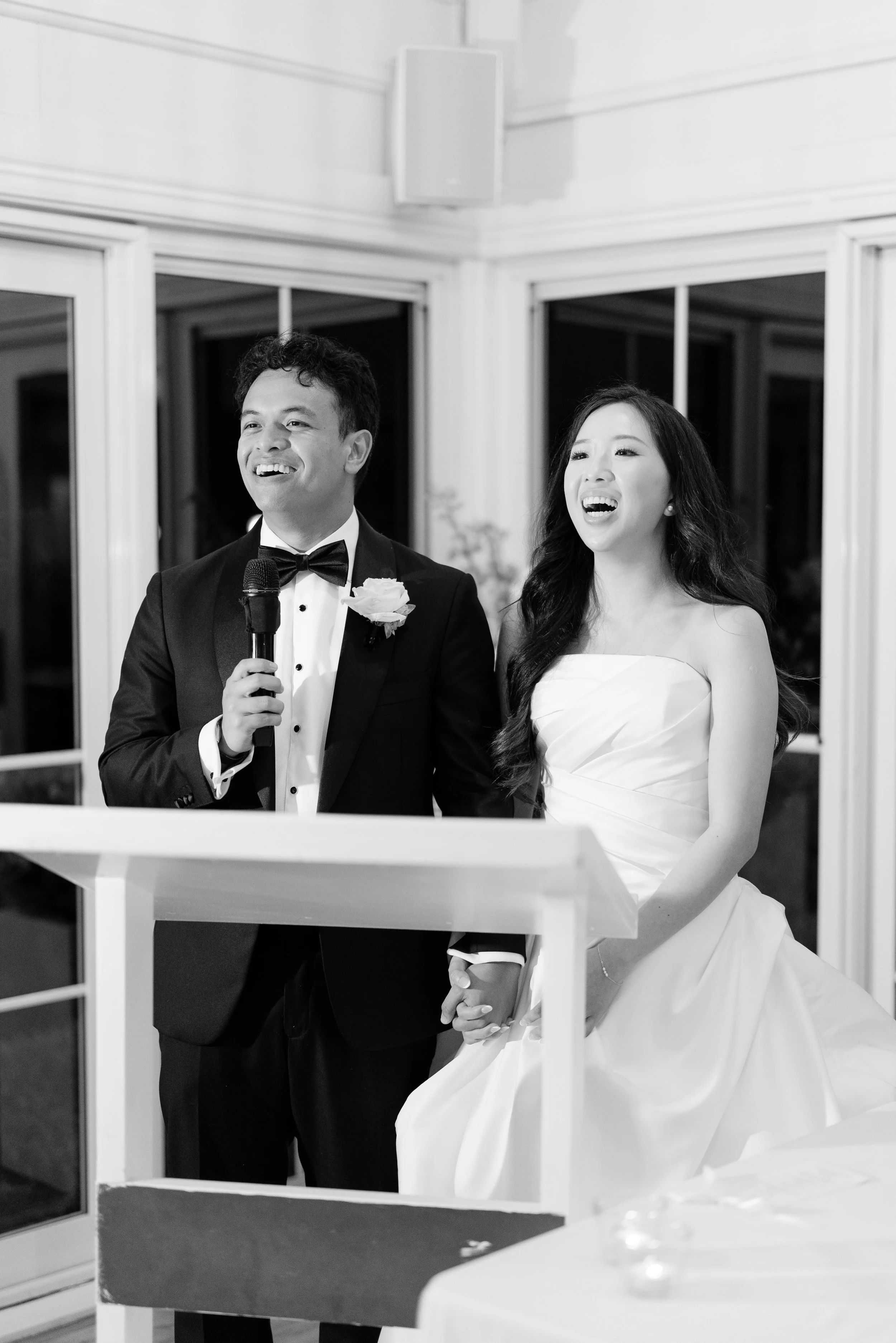 Alicia and Ezra at a microphone, Ezra speaking while Alicia laughs beside him, in black and white