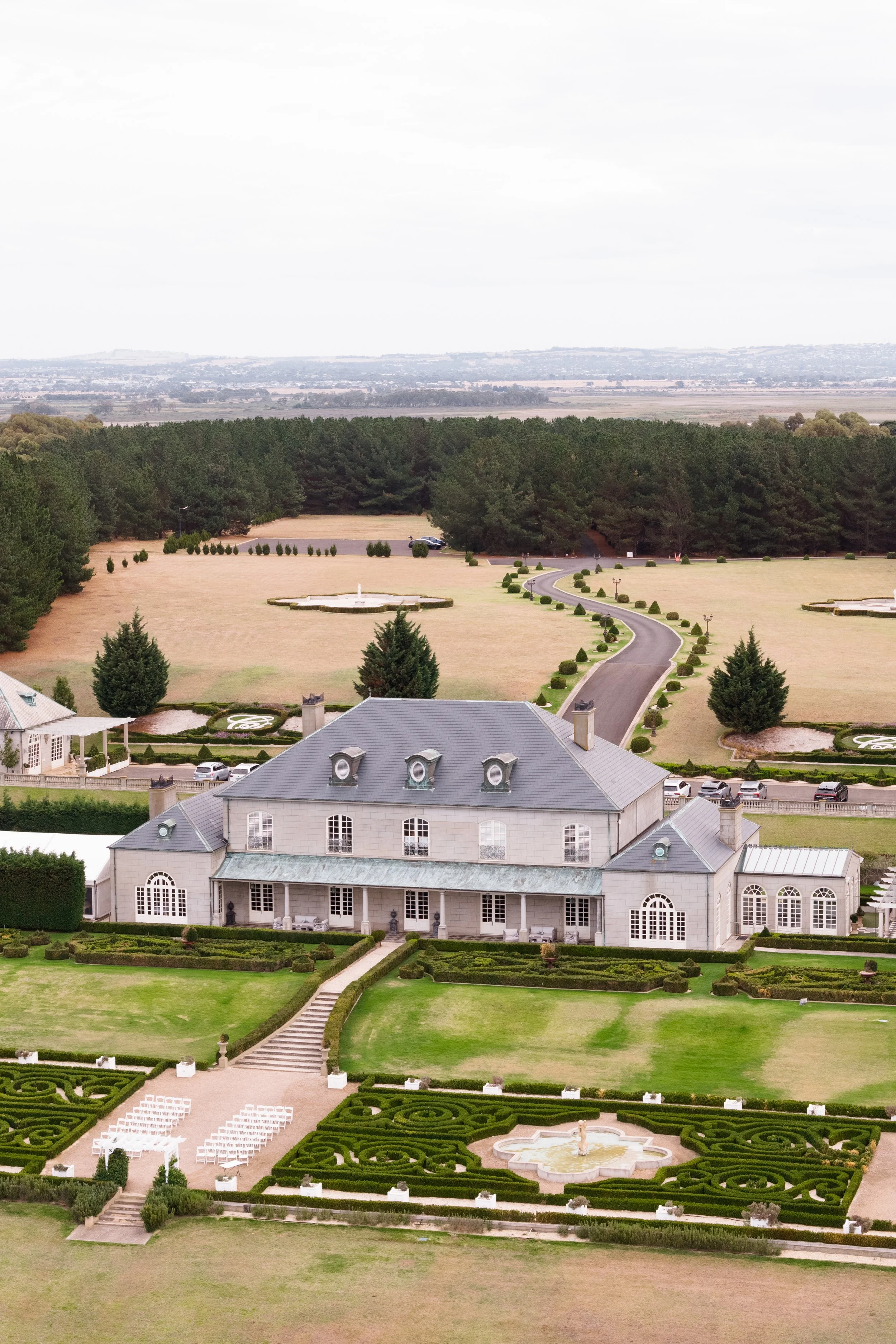 Large estate with a grand gray mansion, manicured garden, fountains, and winding driveway, surrounded by lush trees and open lawns, with a distant landscape under cloudy sky.