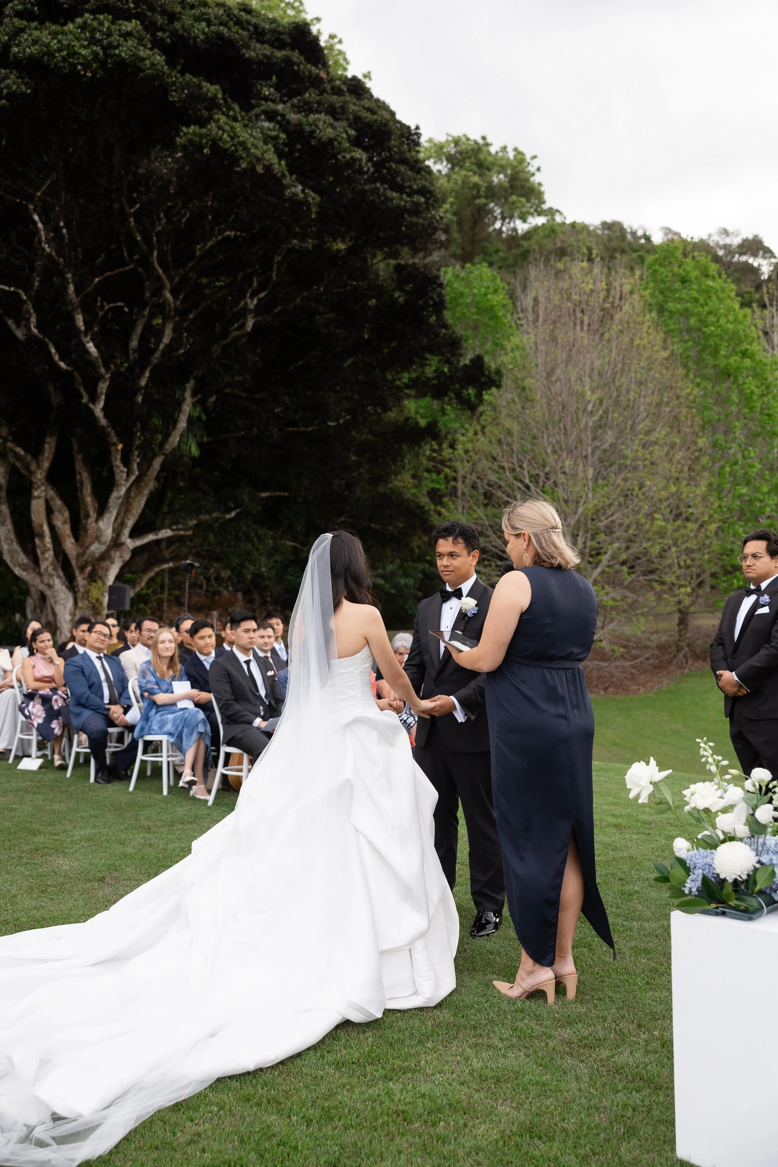 Alicia and Ezra at the celebrant during the outdoor ceremony, guests seated behind her long gown train