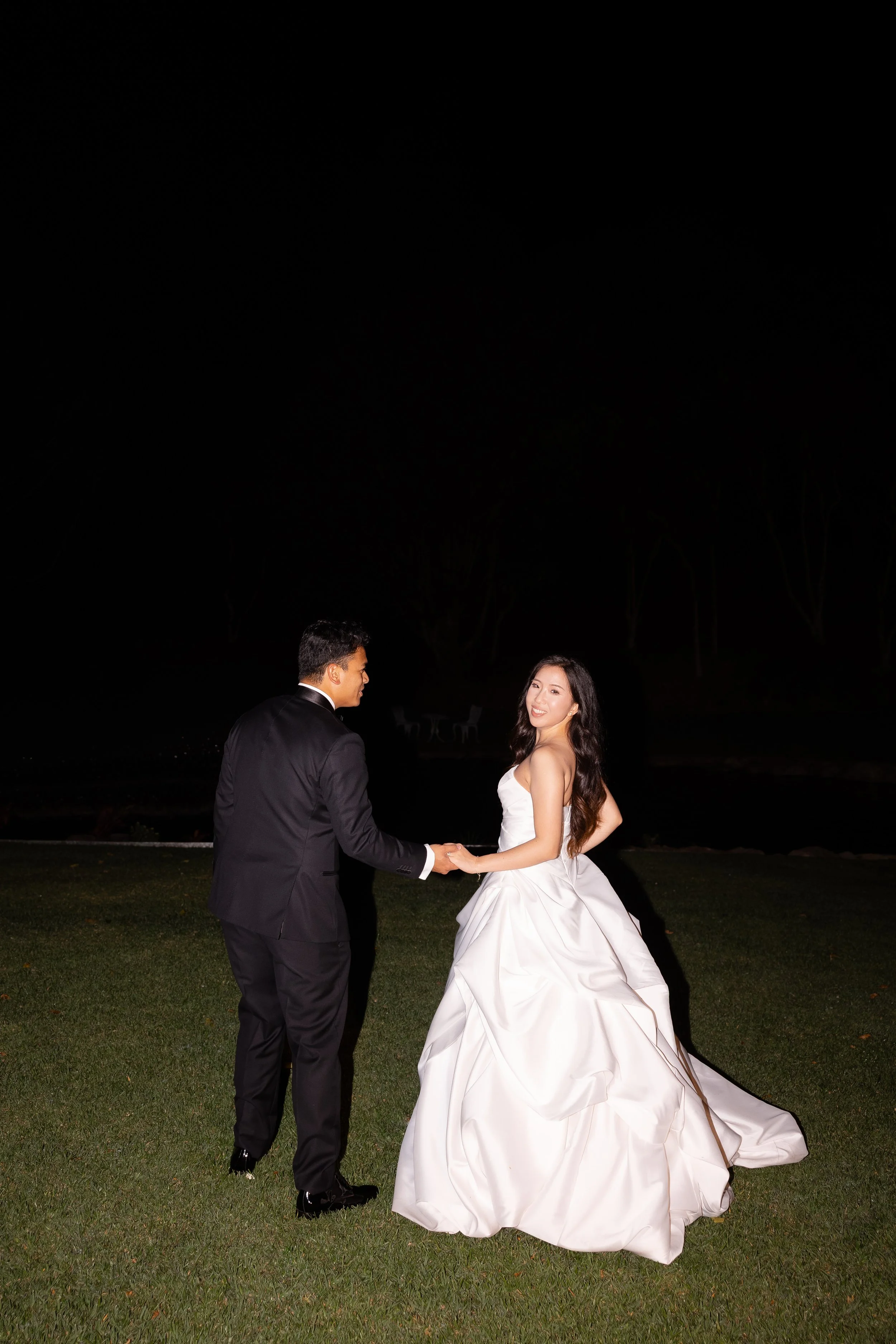 Alicia and Ezra holding hands on a dark lawn at night, Alicia in a white gown looking over her shoulder