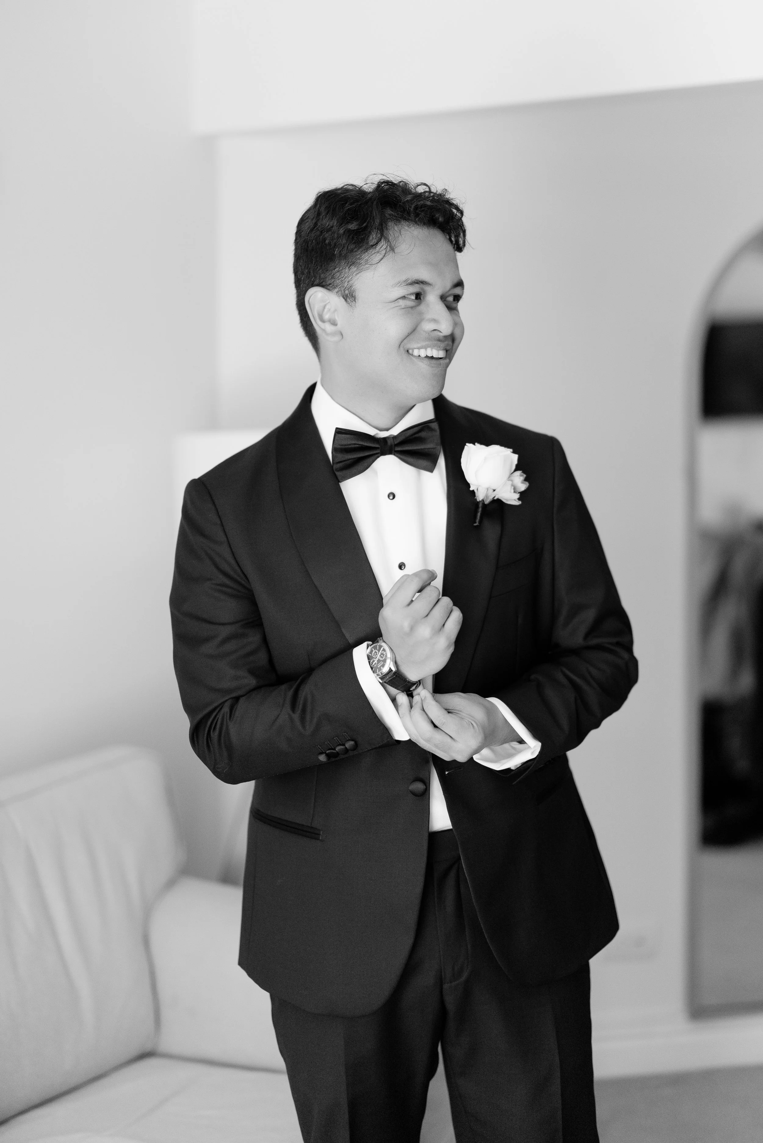 Alicia and Ezra's wedding day: Ezra smiling in a black tuxedo with a white boutonniere, in black and white
