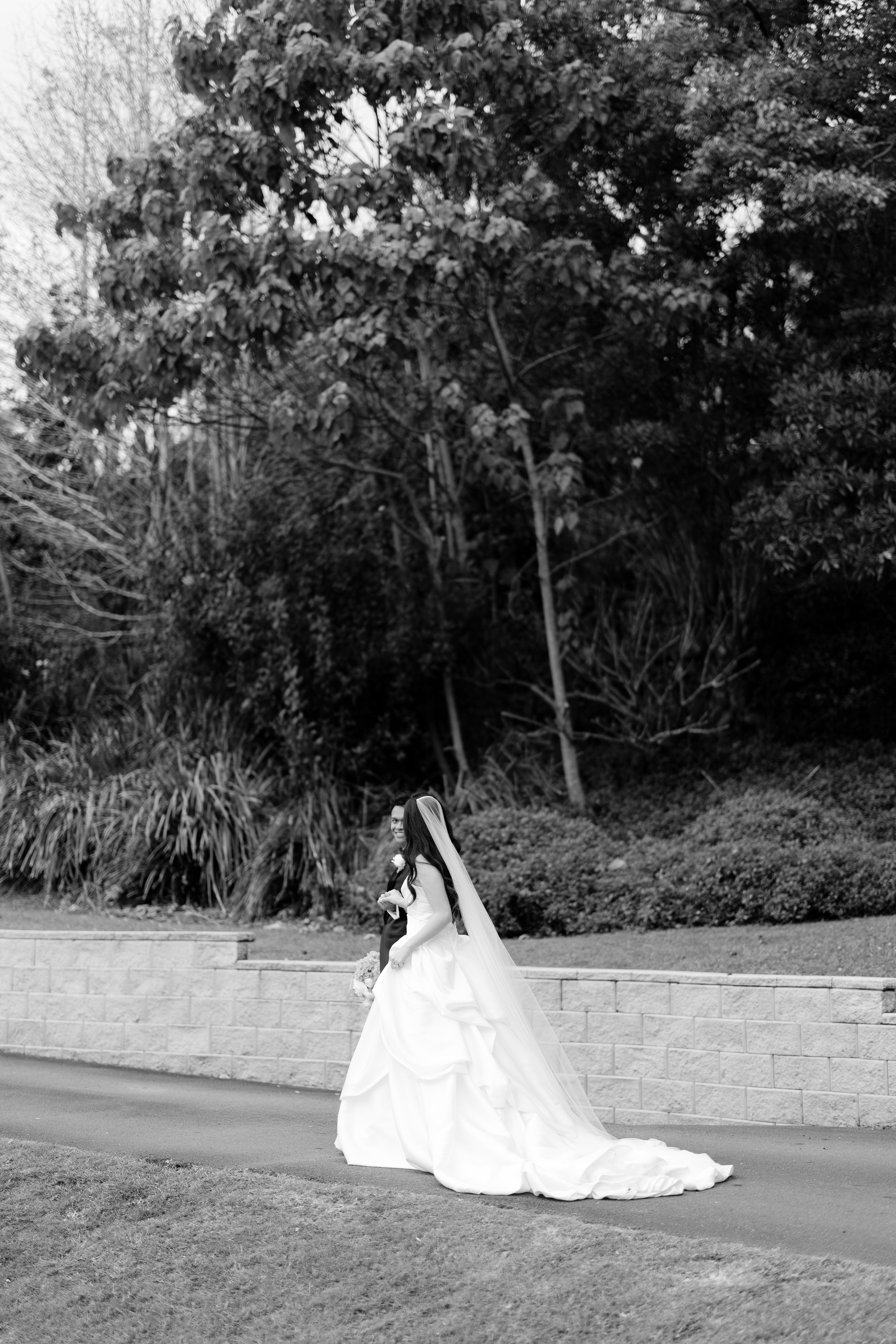 Alicia in her wedding gown and long veil beside a low wall under tall trees, in black and white