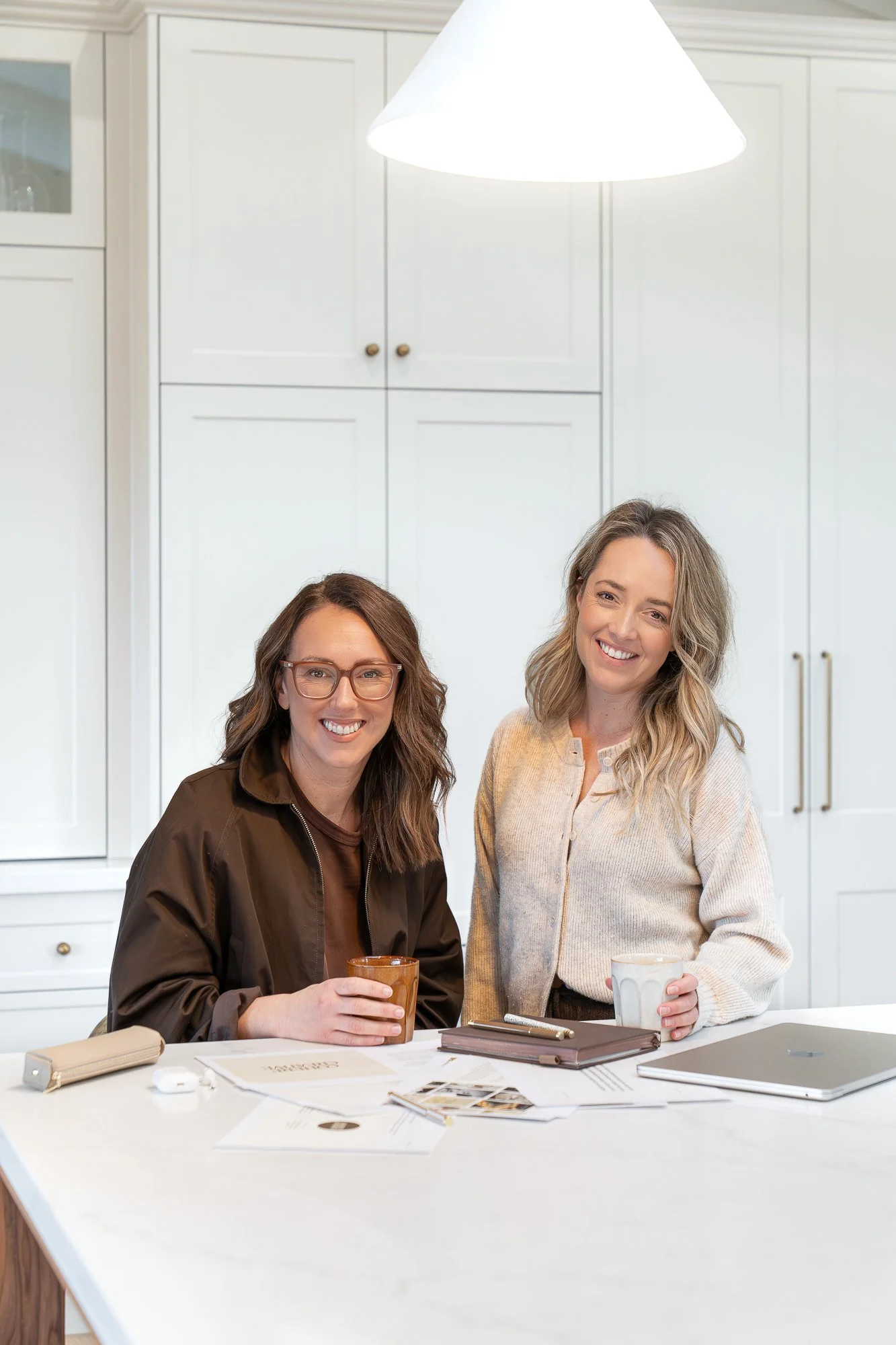 Jess Scholz and Lauren Maxfield standing in a kitchen, smiling and holding coffee mugs, with papers and a tablet on the white kitchen counter in front of them.