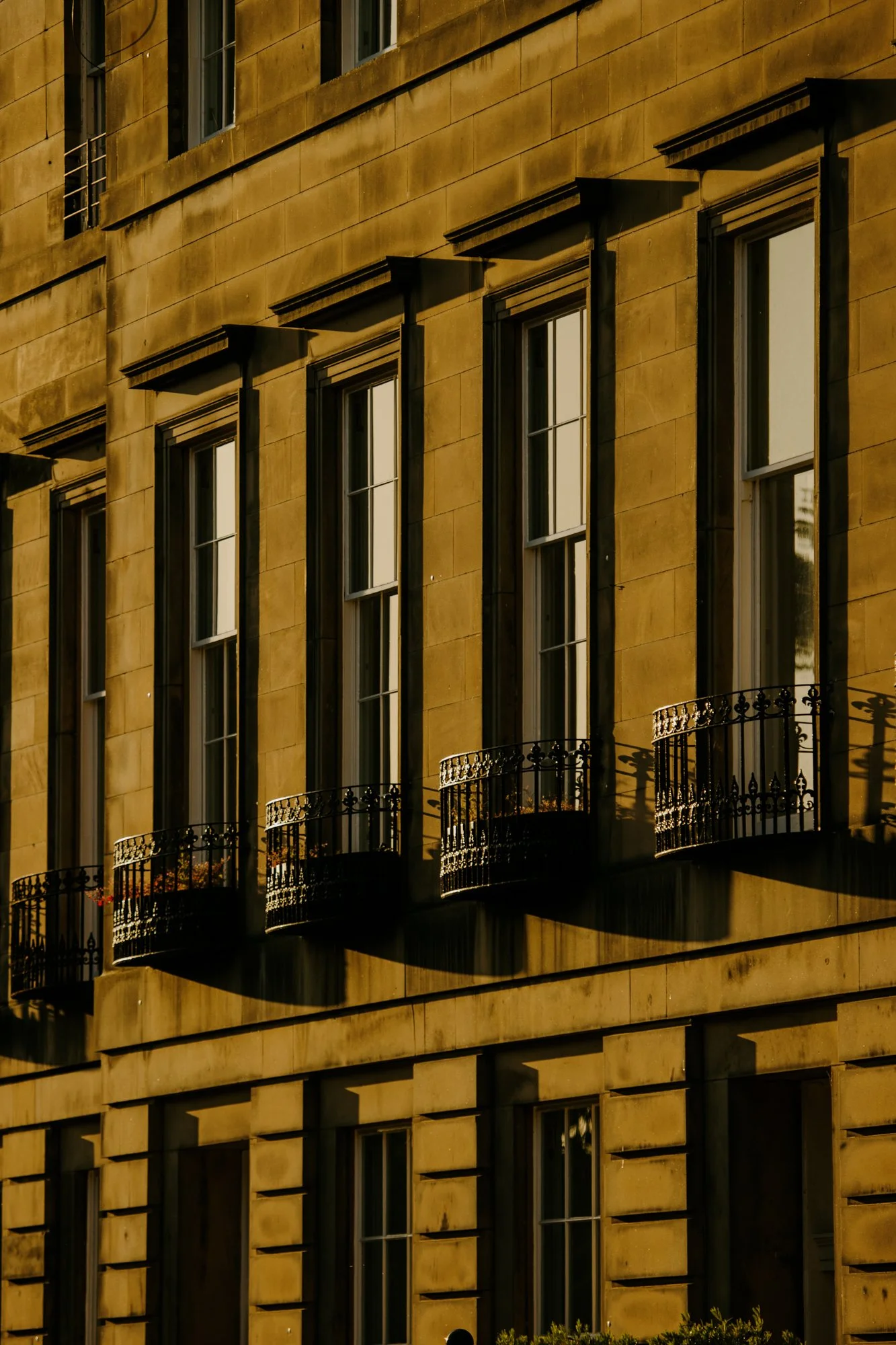 A close-up view of the exterior of an Edinburgh townhouse with multiple tall windows, some with small black iron balconies, casting shadows on the wall.