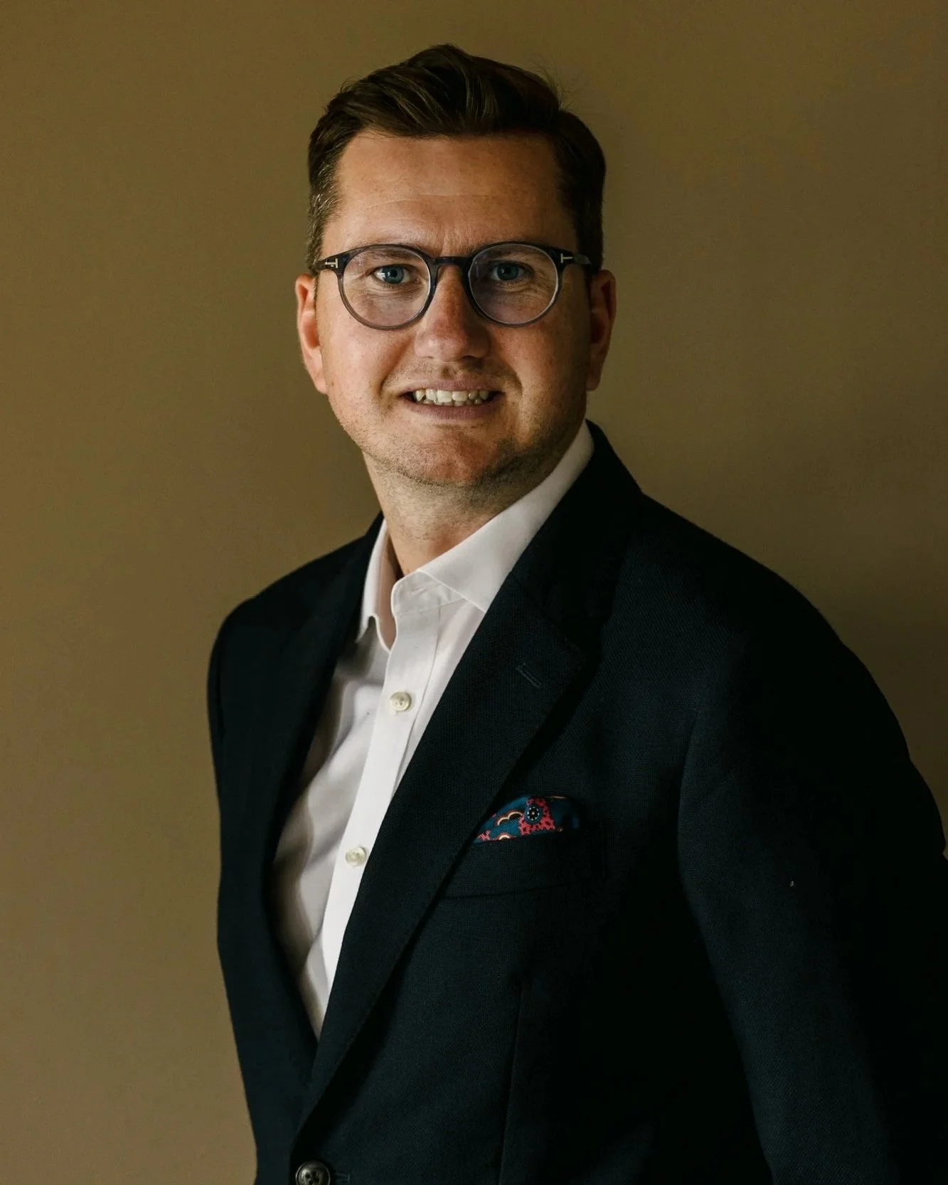 Jamie McNeill wearing glasses, a suit, and a white shirt, smiling and looking at the camera.