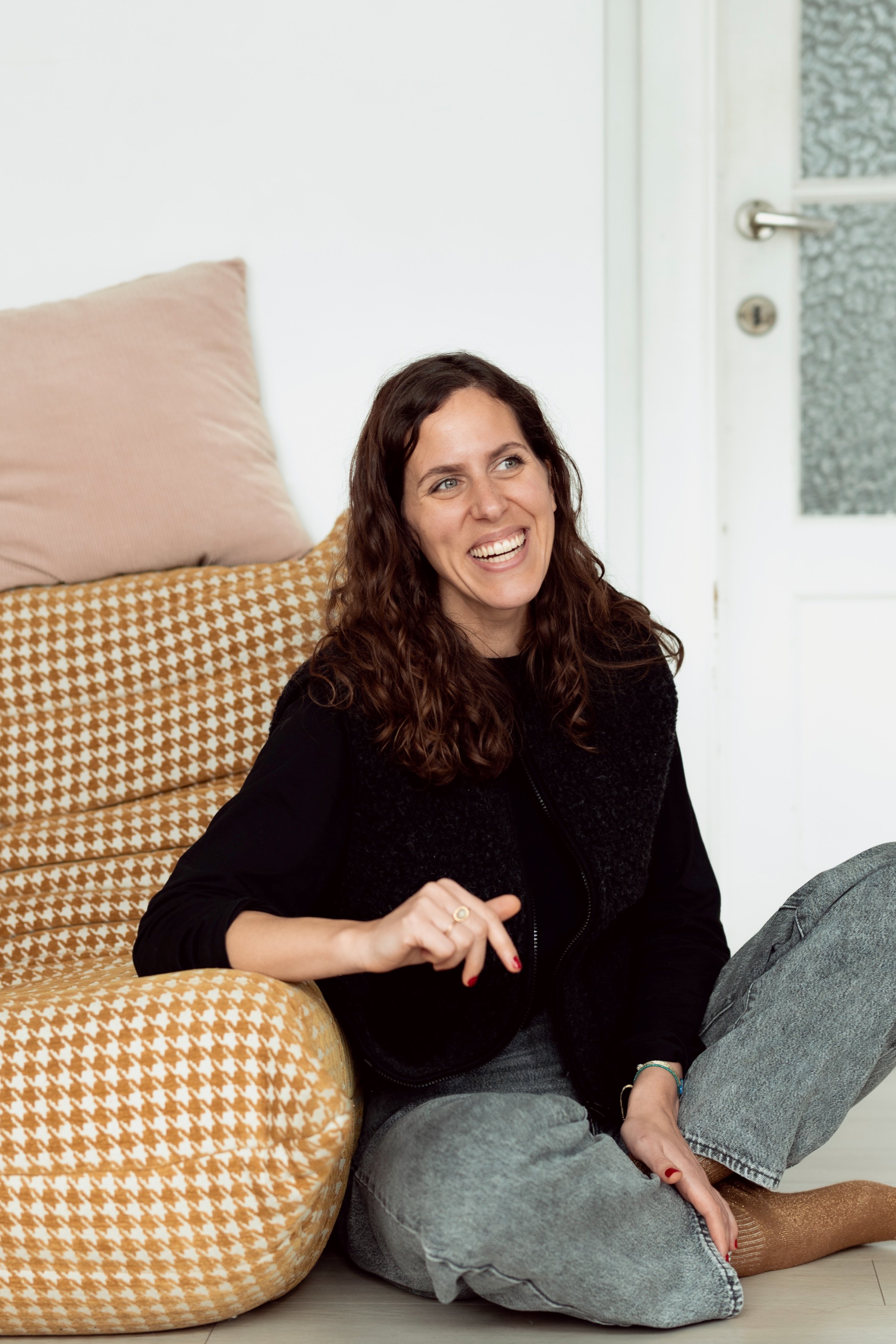 Une femme aux cheveux bouclés bruns souriante, assise par terre à côté d'un fauteuil orange à motif pied-de-poules, dans une pièce lumineuse avec une porte blanche en arrière-plan.