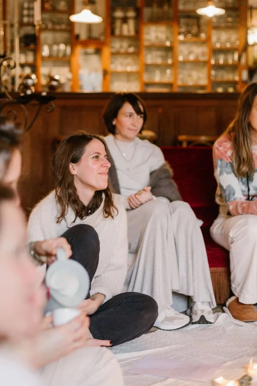 Groupe de personnes assises dans une pièce chaleureuse, discutant ou écoutant attentivement, avec étagères remplies d'objets en arrière-plan.