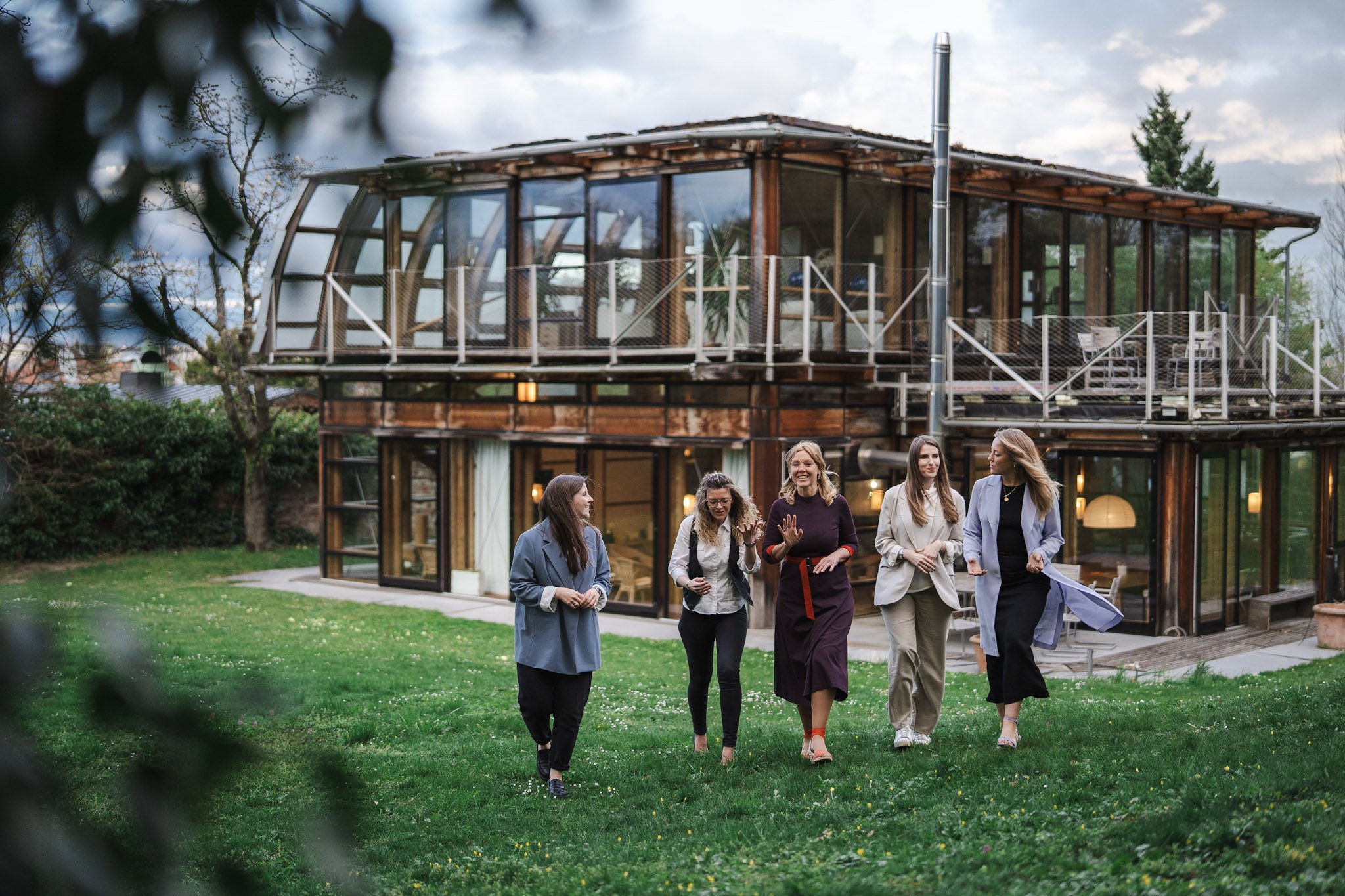 A group of six women walking and talking outside near a modern glass and wood house, with a tree and cloudy sky in the background.
