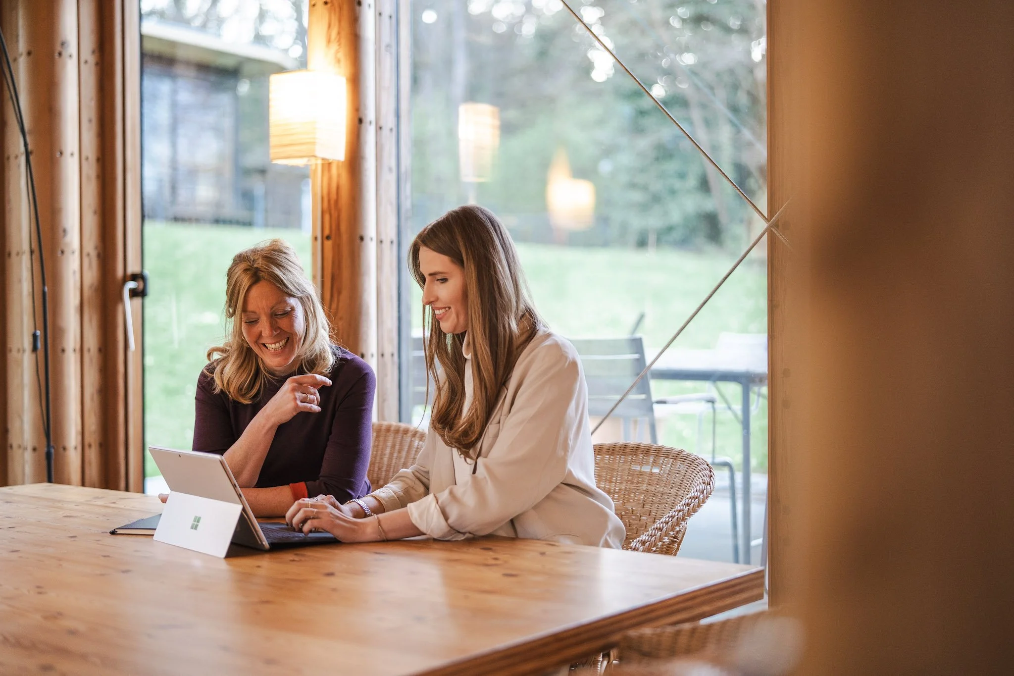 Two women sitting at a wooden table, looking at a tablet together in a bright room with large windows and greenery outside.