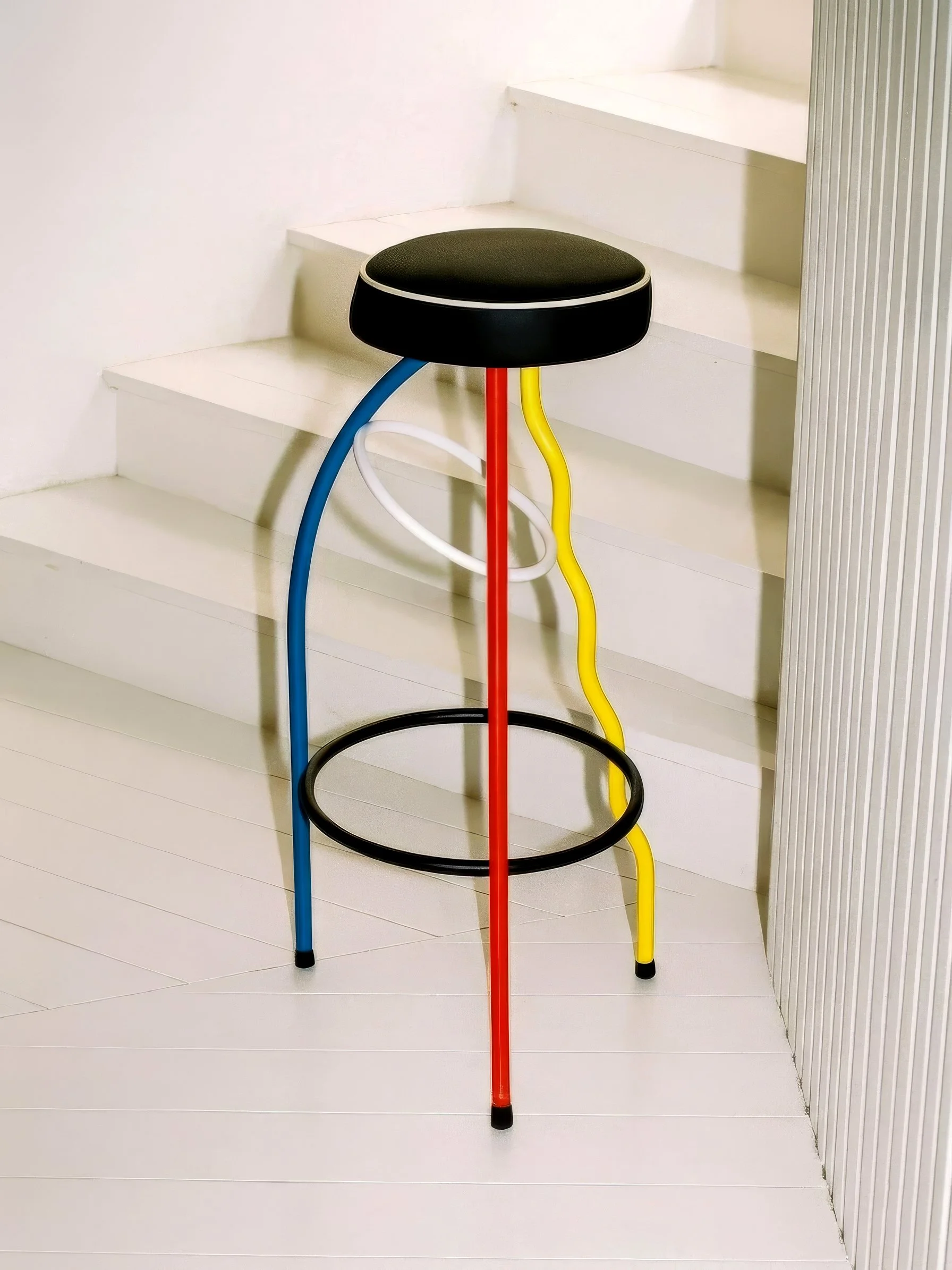 Tabuerte Dúplex, pieza postmoderna diseño icónico Español. Con estructura de metal multicolor, con patas en azul, rojo, amarillo y negro, y asiento redondo negro, colocada junto a una escalera blanca y pared blanca.