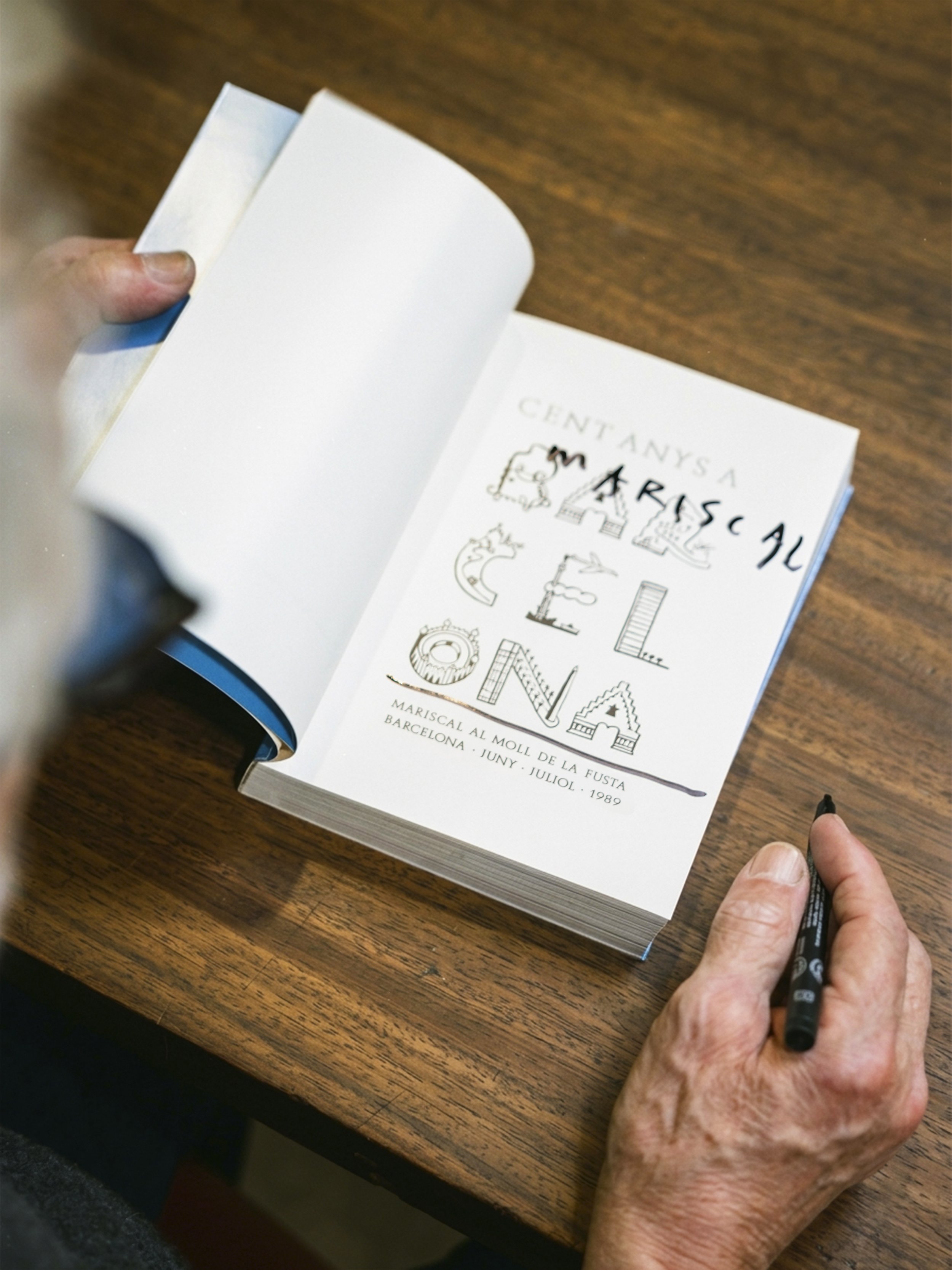 Javier Mariscal firmando y dedicando su libro Traveling Book Los Ángeles, publicado por Louis Vuitton. Escribe con letras grandes y gestuales, sentado en una mesa blanca