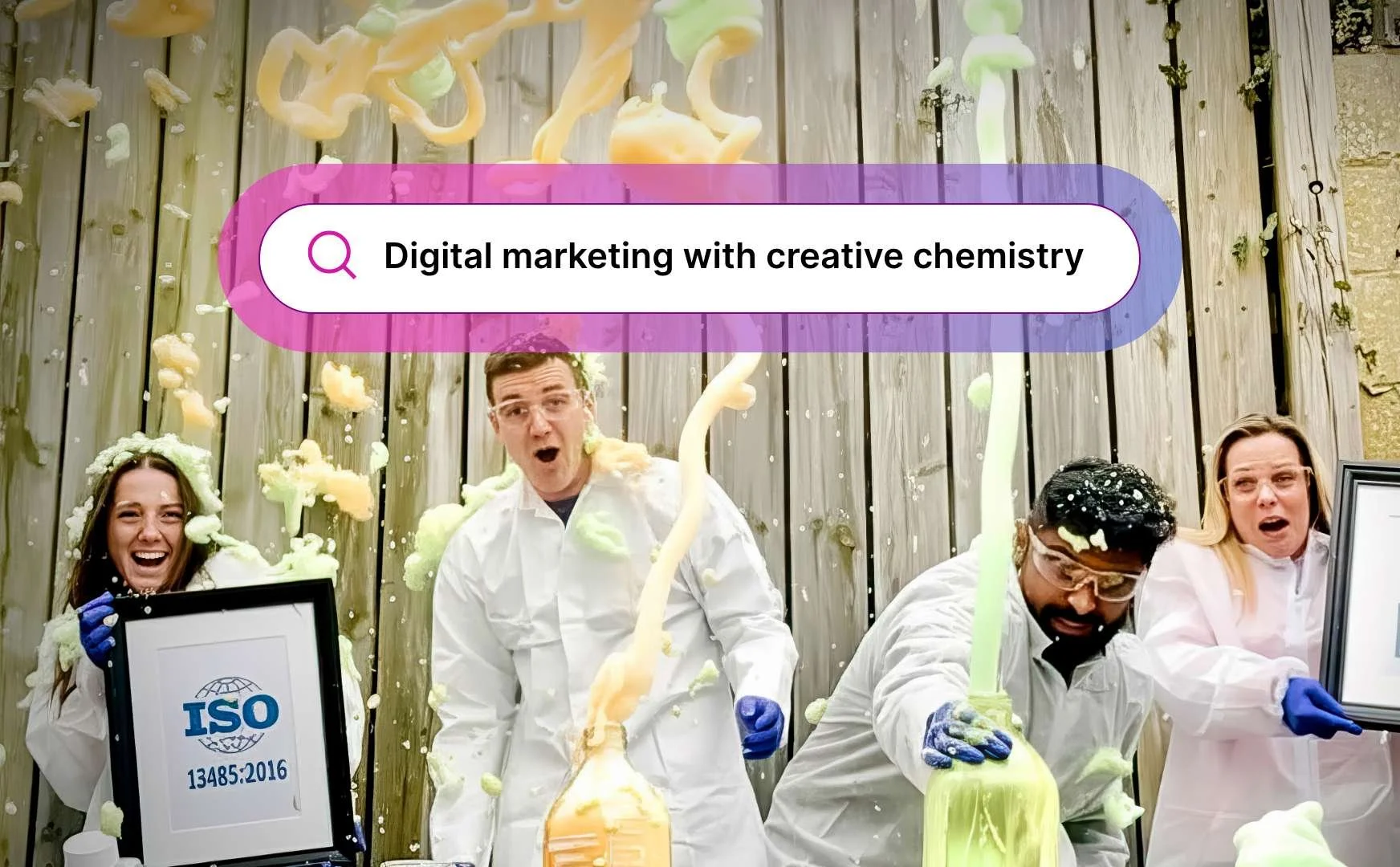 Group of four diverse people dressed in white lab coats and safety goggles engaging in a colorful foam experiment outdoors, with a wooden fence background, a framed ISO certificate, and a search bar graphic that reads 'Digital marketing with creative chemistry'.