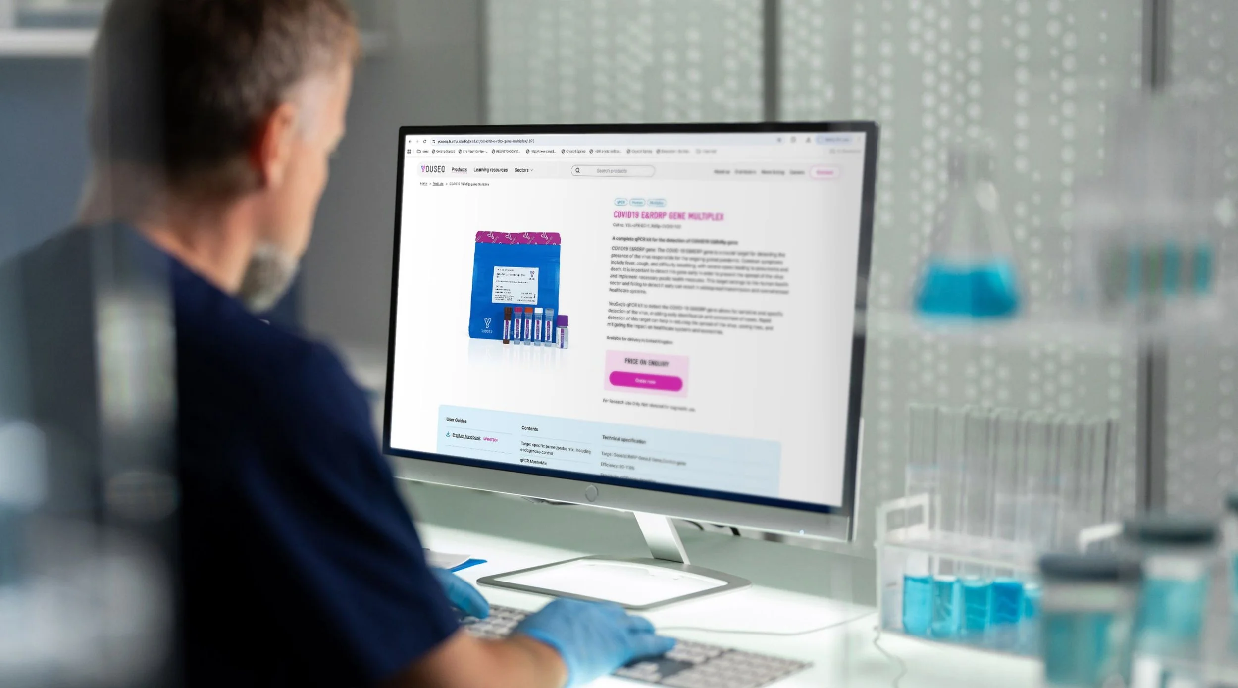 A person with gray hair wearing a face mask and blue gloves looking at a computer monitor displaying a product page for COVID-19 test kits.