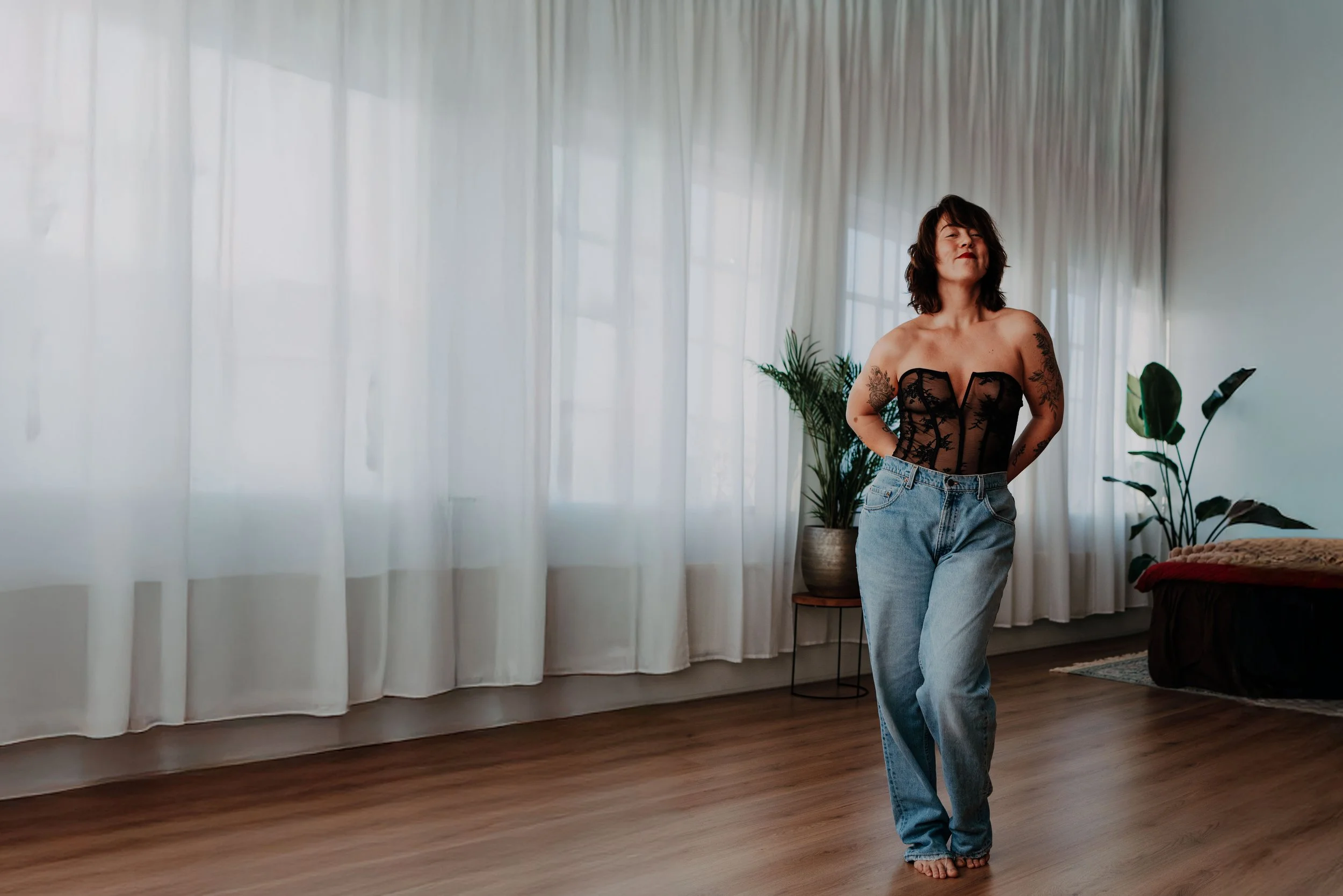 Vrouw met schouderlange haar in een luide top en lage blauwe jeans, staand in een lichte woonruimte met witte gordijnen en planten.
