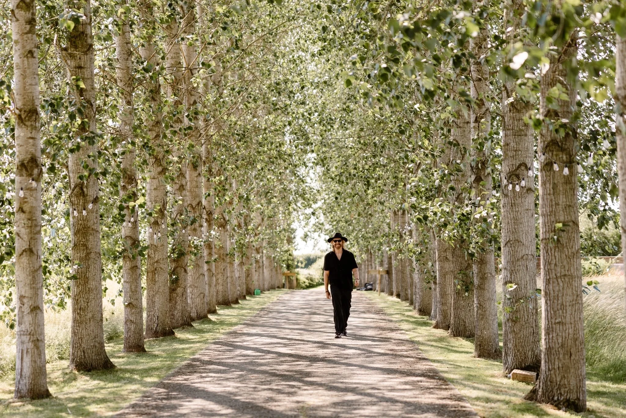 Alternative wedding singer at a beautiful UK wedding venue walking down a beautiful avenue of trees with dappled light beams through the leaves