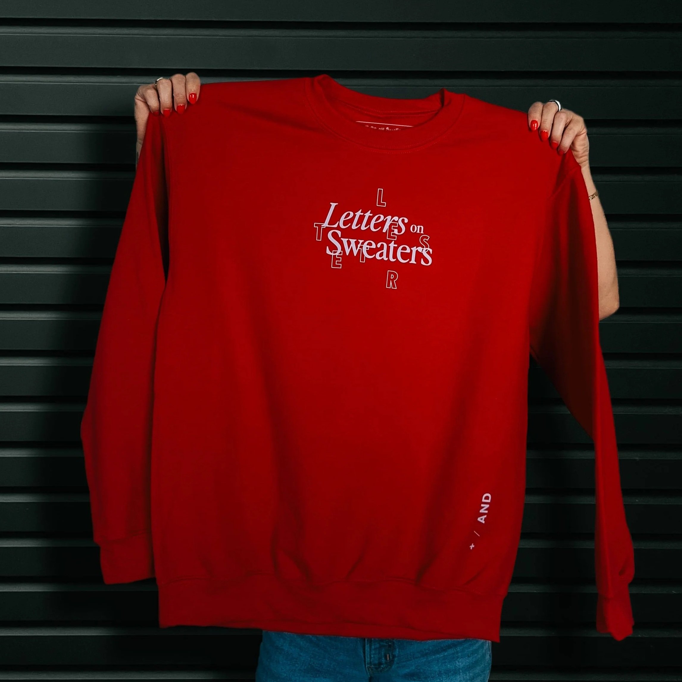 Holding a cherry red crewneck sweatshirt that has Letters on Sweaters designed on it in lavender letters