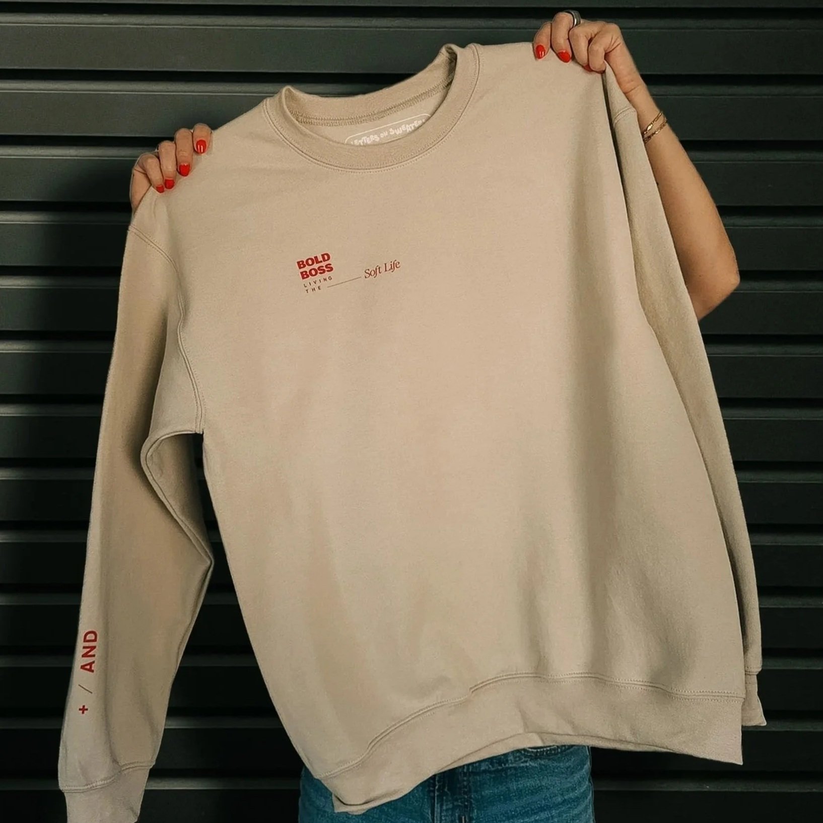 Holding sand colored sweatshirt with small design that says Bold boss living the soft life.
