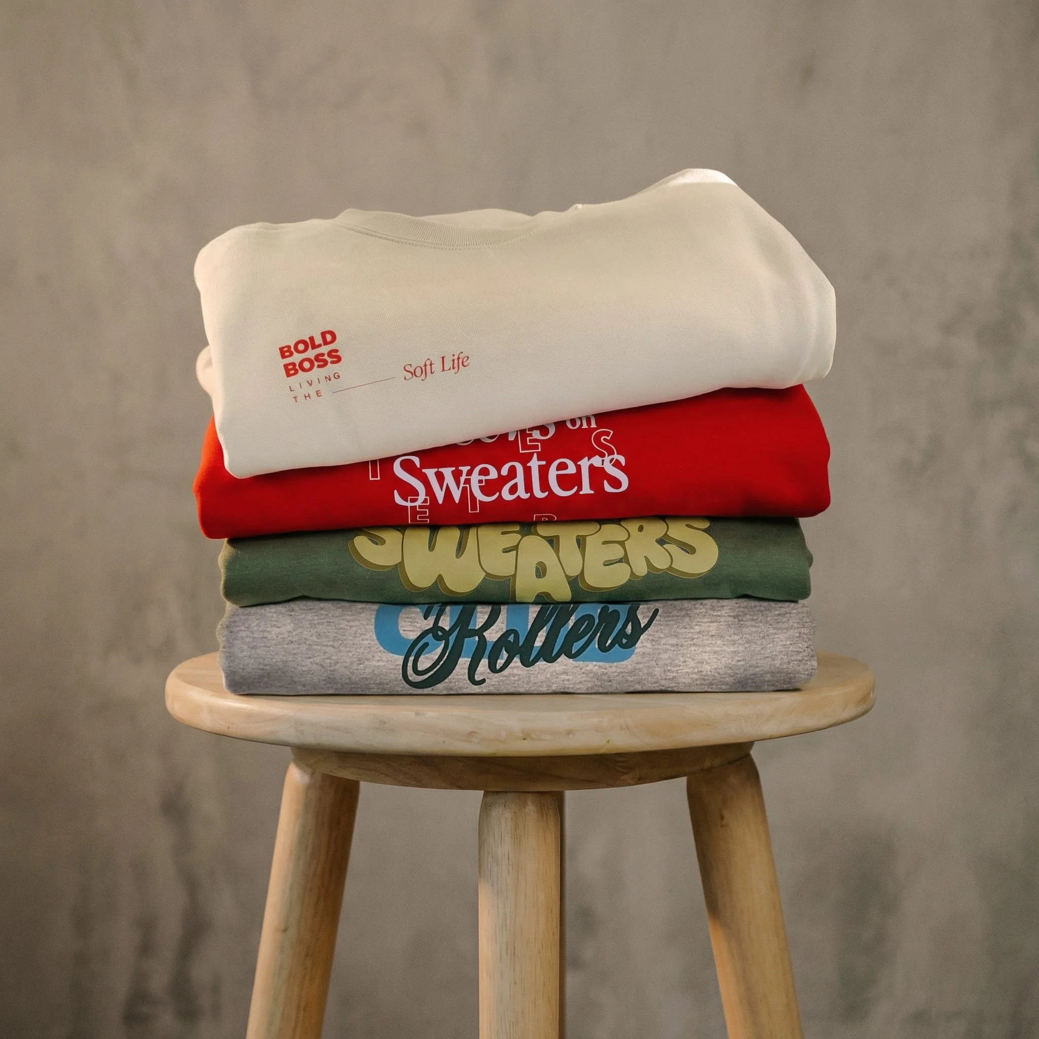 4 crewneck sweaters designed by Letters on Sweaters folded on a wood stool.