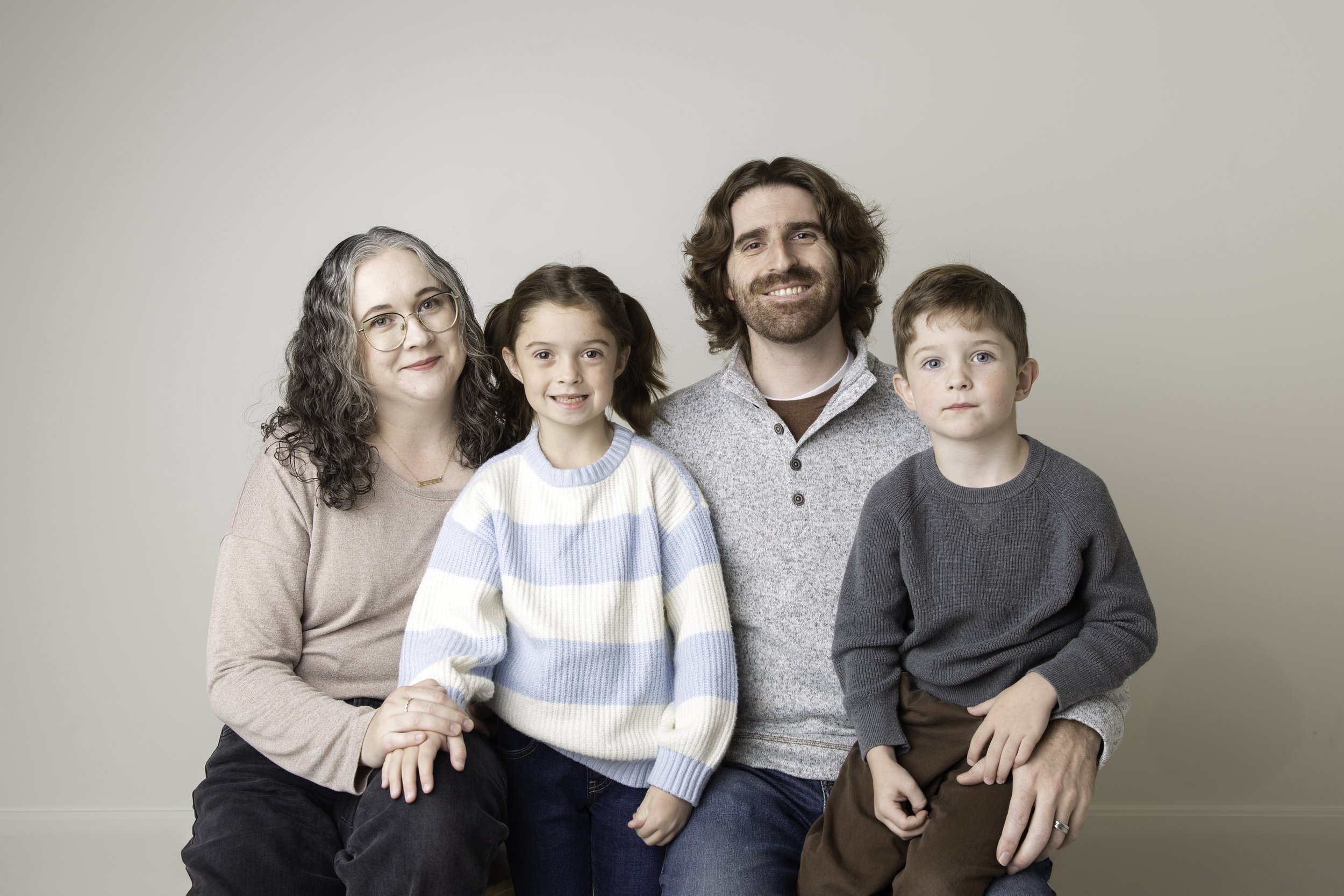 a family with a mom and dad and two children sitting together smiling against a white wall during a studio photo session