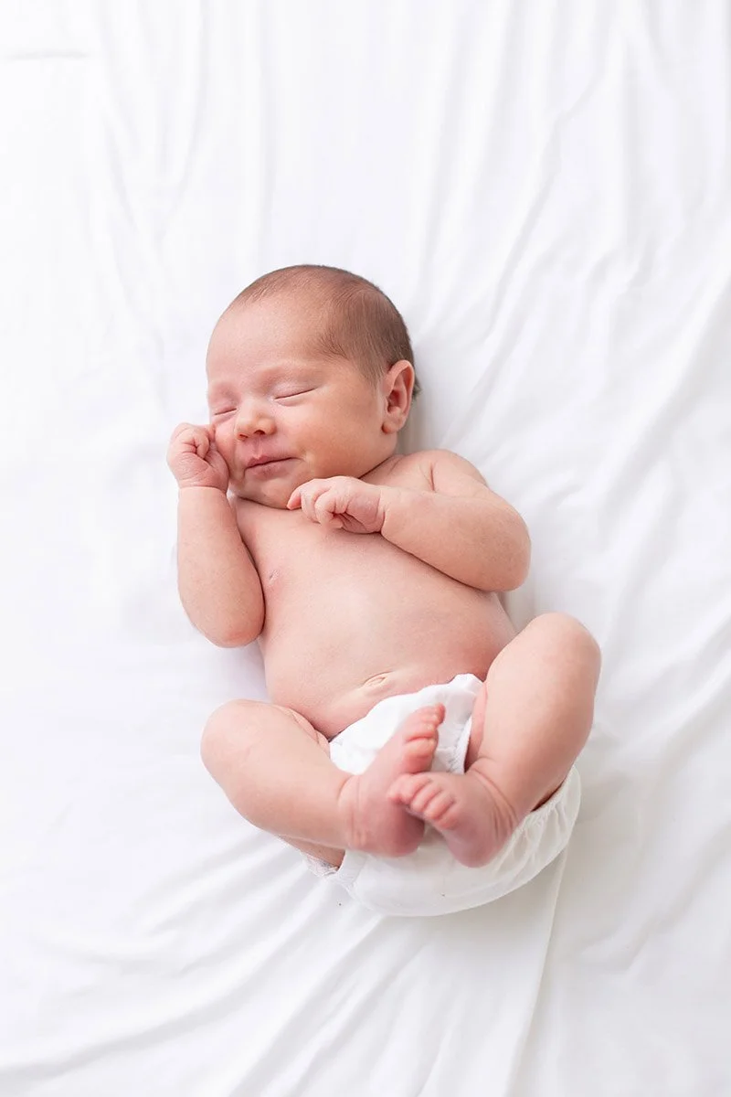 A sleeping newborn baby lying on a white blanket, wearing a diaper with one hand near the face and the other resting on the chest.