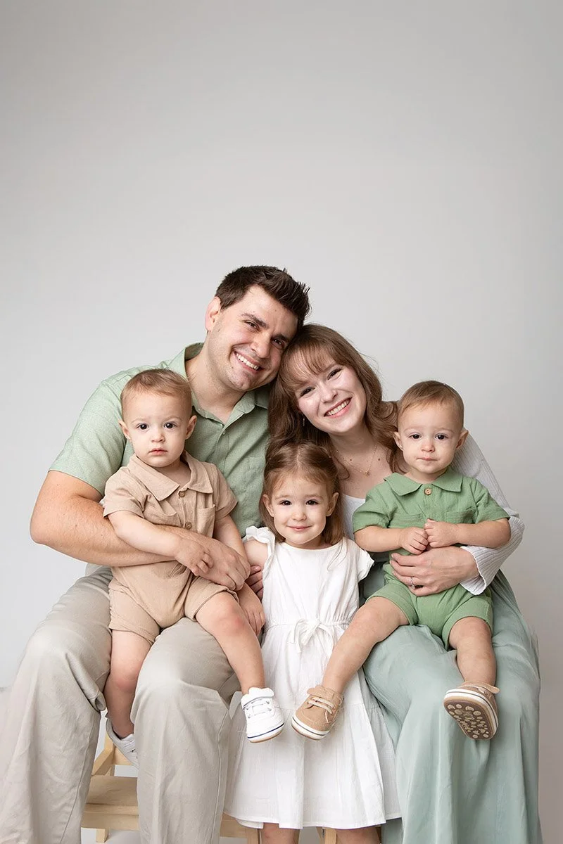 Family of five sitting together and smiling, including a father, mother, and three young children, against a plain light background.