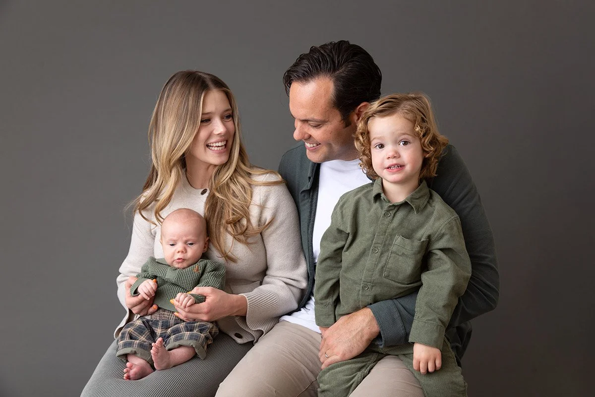 A family of four sitting together against a plain gray background, including a woman holding a baby, a man, and a young boy sitting on the man's lap.