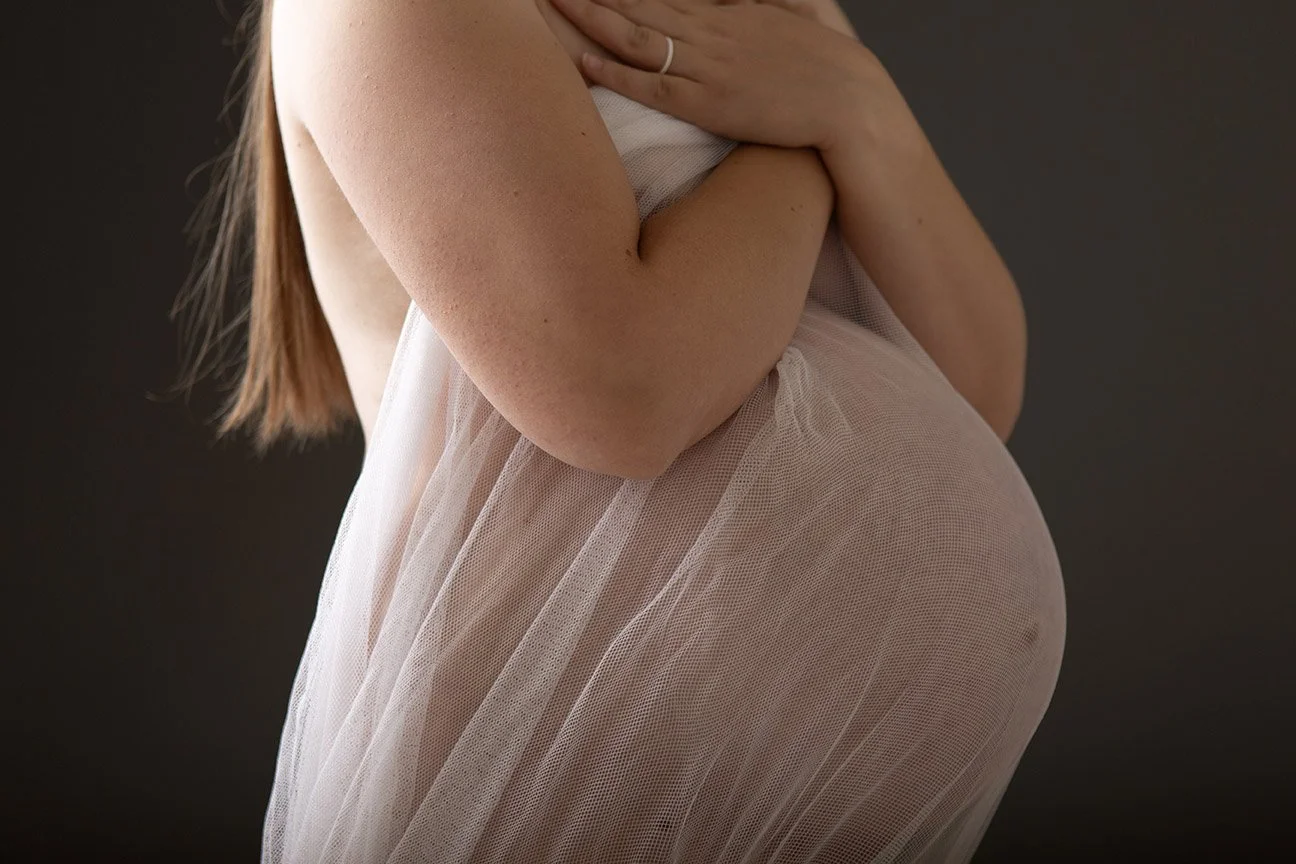 Close-up maternity portrait showing a pregnant woman’s baby bump wrapped in soft sheer fabric for a fine art pregnancy photograph