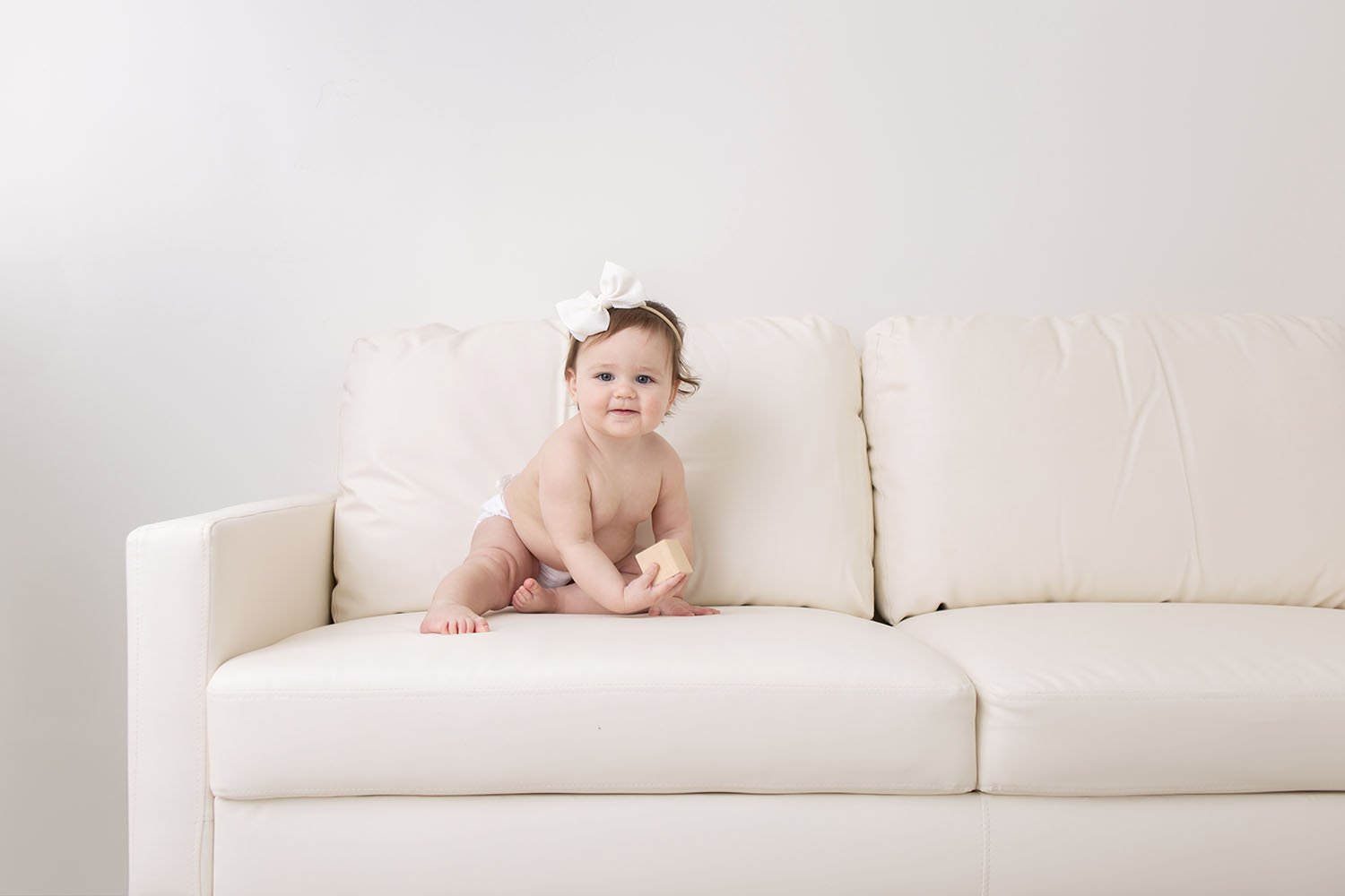 one year old baby holding a wooden block while sitting on a white sofa during a studio photo session