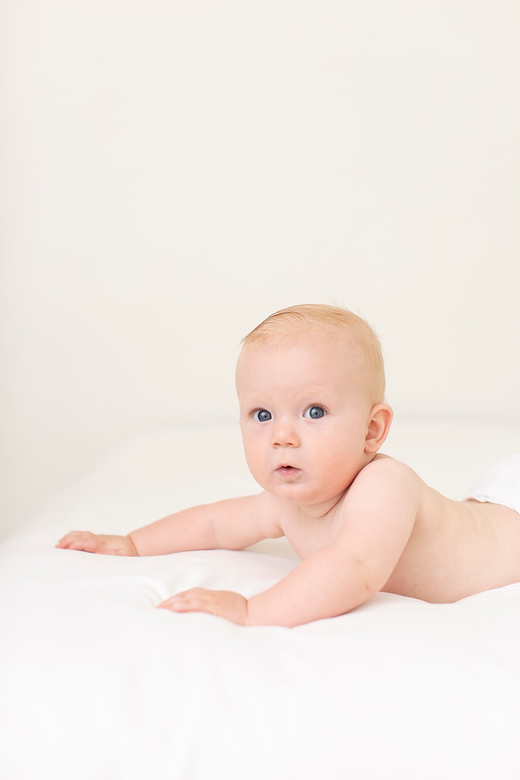 A baby with blue eyes and light hair lying on a white surface, looking at the camera.