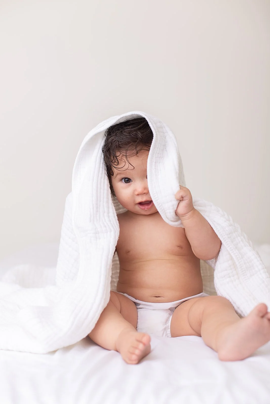 Smiling baby sitting on a white bed with a blanket draped overhead during a bright lifestyle studio session