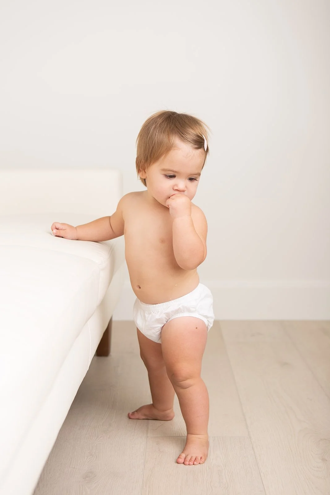 A young toddler standing barefoot near a white couch, wearing only a diaper, with one hand on the couch and the other hand in their mouth, looking down.