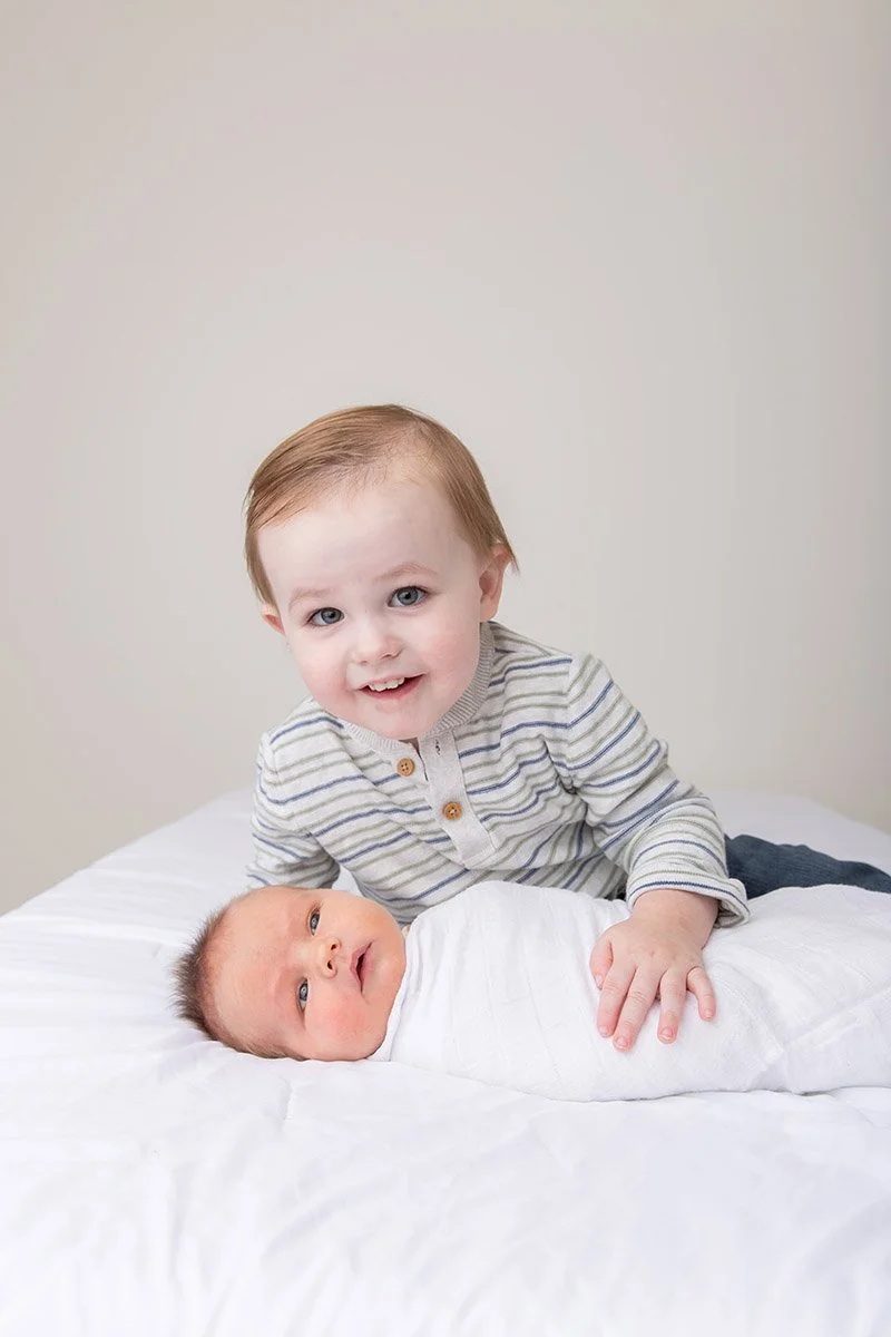 Young boy with light brown hair and blue eyes smiling while leaning on a newborn baby wrapped in a white blanket, lying on a white bed, against a plain beige wall.