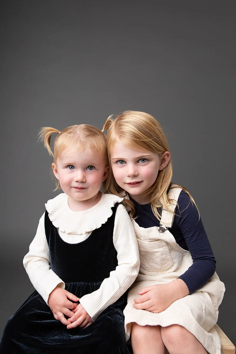 Two young girls sitting closely together against a gray studio background.