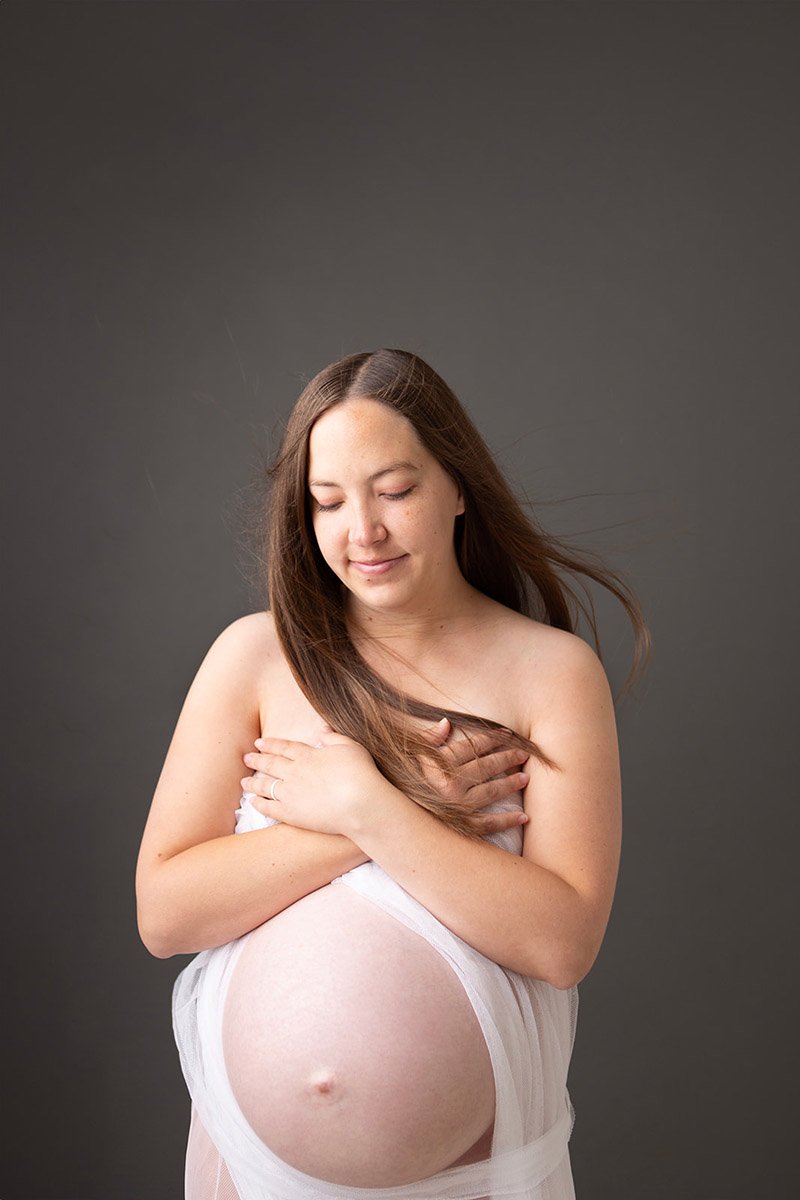 A pregnant woman with long brown hair, eyes closed, smiling gently, holding her baby bump with both hands, wearing a white strapless dress against a gray background.