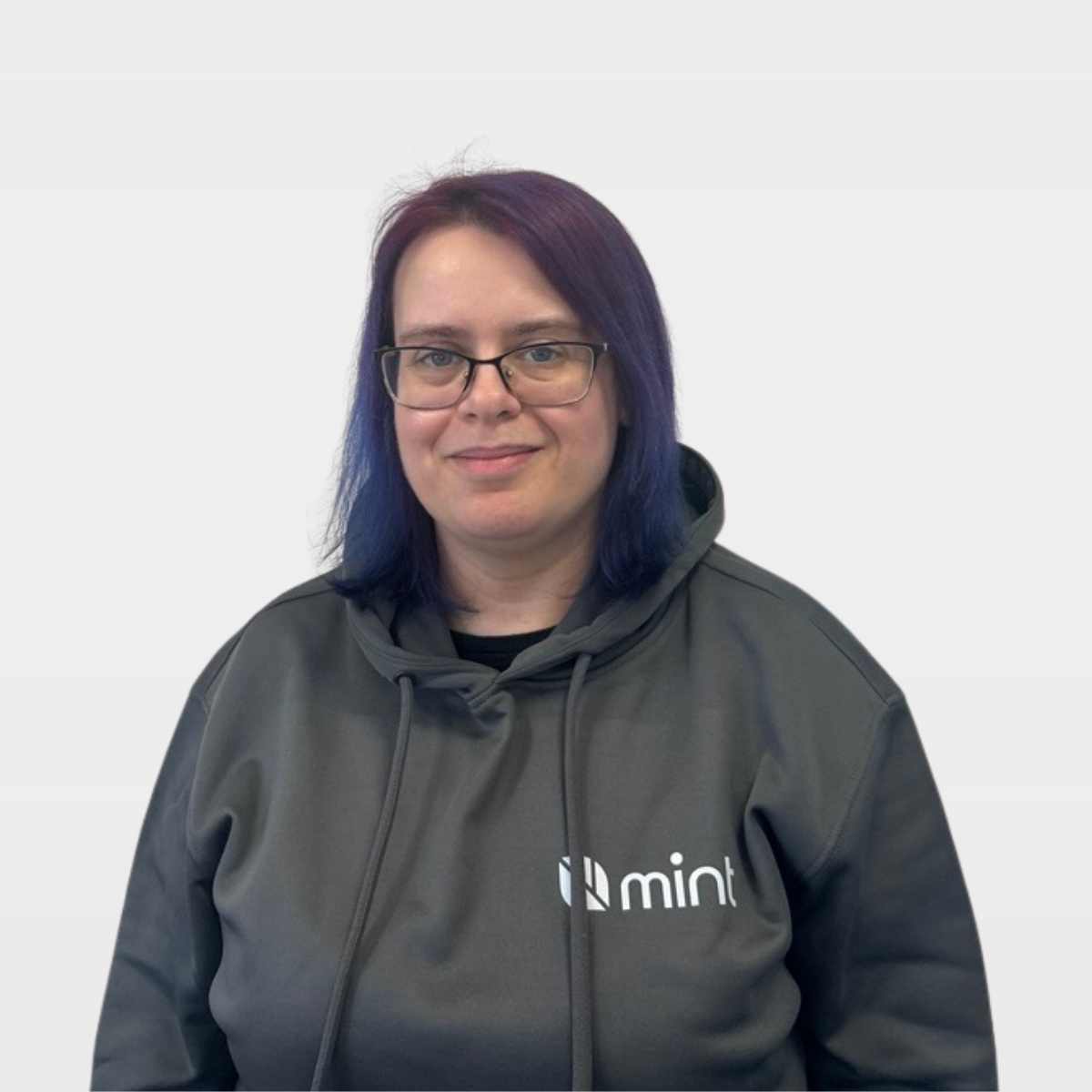 A smiling woman with short dark hair, glasses, and a black top with colorful patterns, standing against a plain white wall.