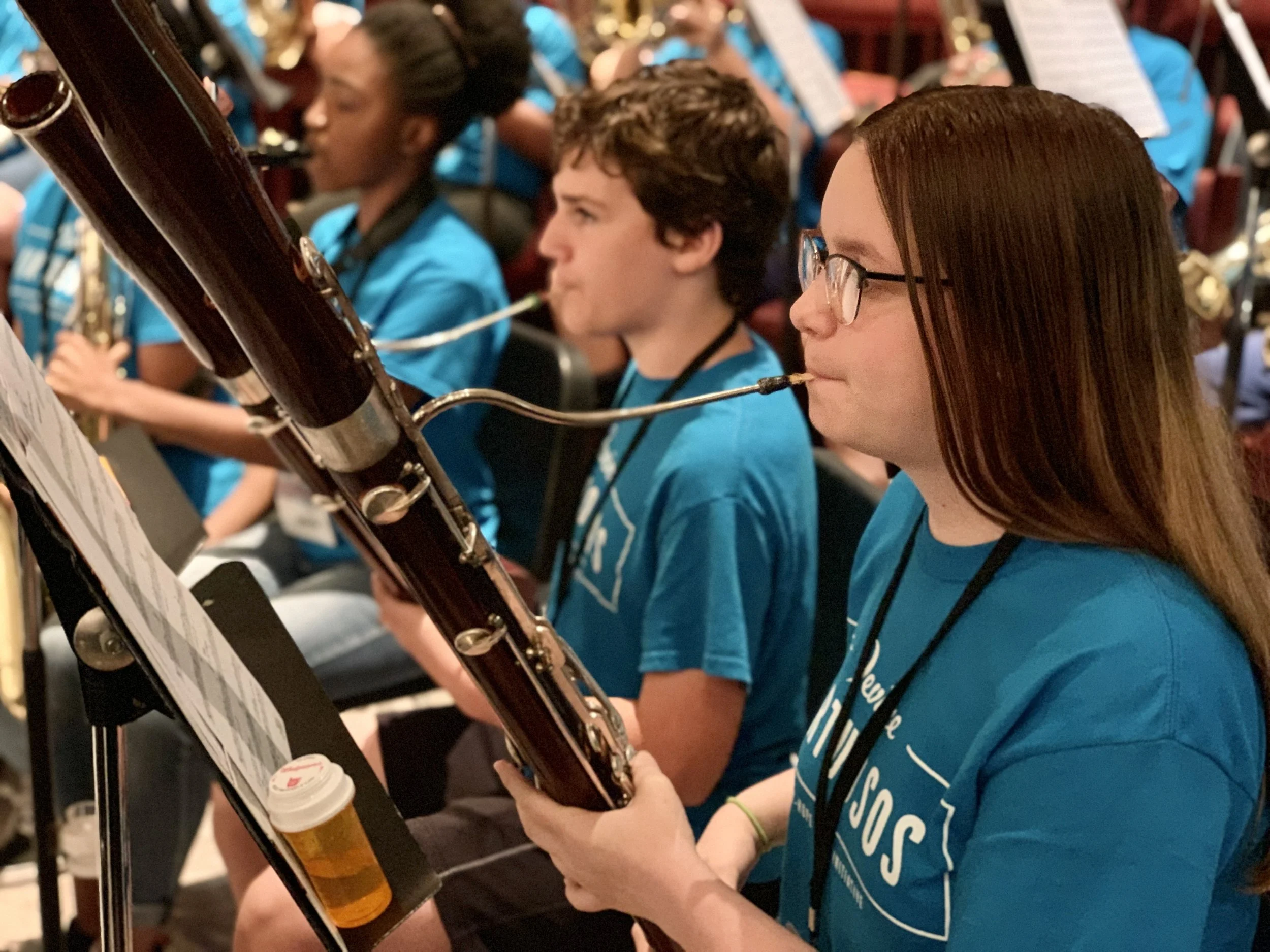 Close-up of young people in blue t-shirts playing bassoon at a music rehearsal, with sheet music on stand and a bottle resting on the stand.