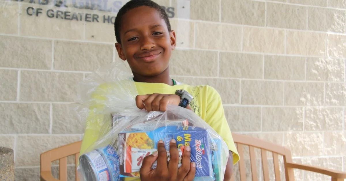 A smiling, young elementary school student holds a clear bag of pantry staple food items from the Houston Food Bank Backpack Buddies Program