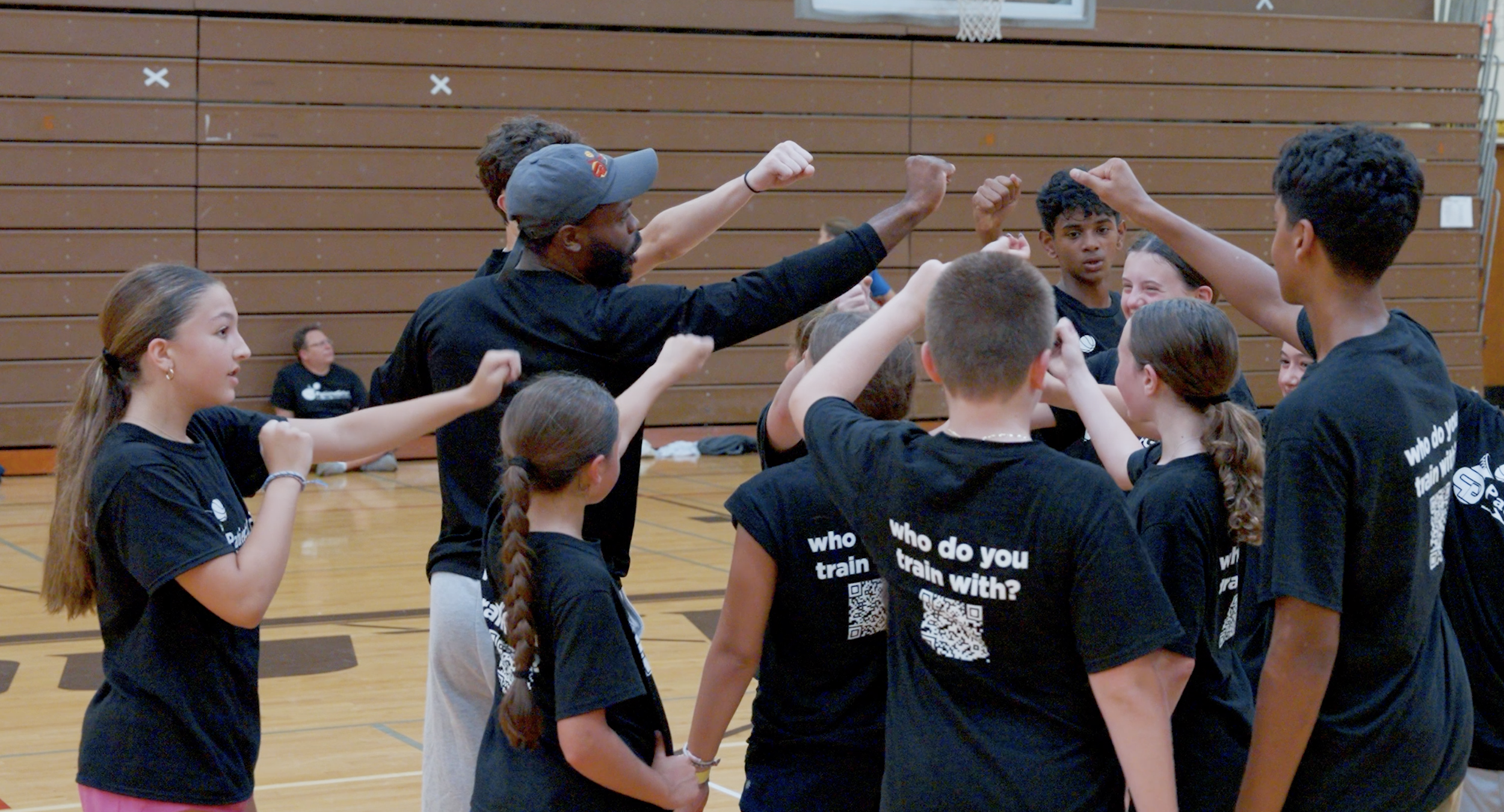 A group of children and an adult coach in a gymnasium, with all participants wearing black shirts that say 'who do you train with?' and raising their fists in a team huddle.
